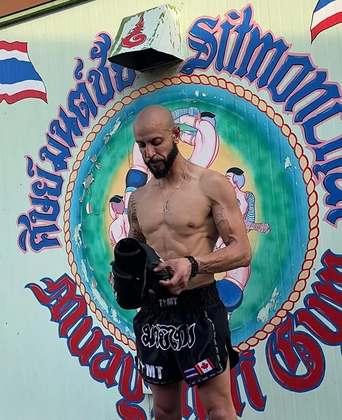 A shirtless man wearing black shorts with white and red Canadian and Thai flags stands in front of a colorful mural with Thai script and fighter illustrations, holding a black item in his hands.
