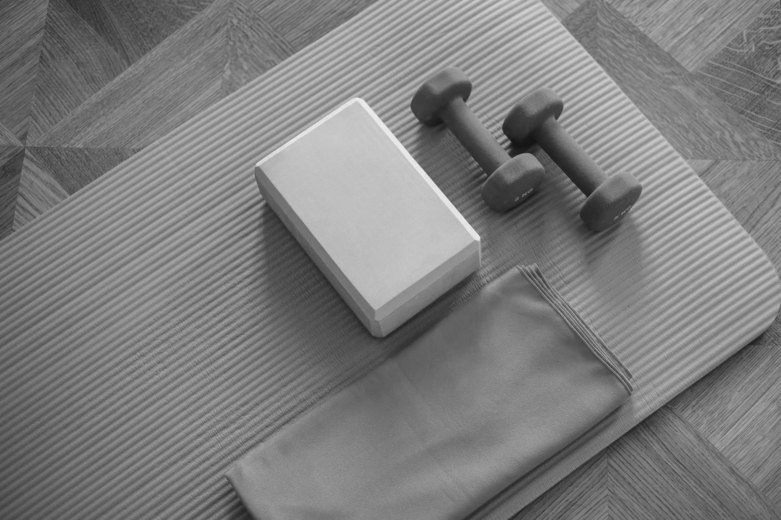 Flat lay of a yoga mat, a pair of dumbbells, a hand grip strengthener, a foam roller, and a carrying case on a wooden floor.
