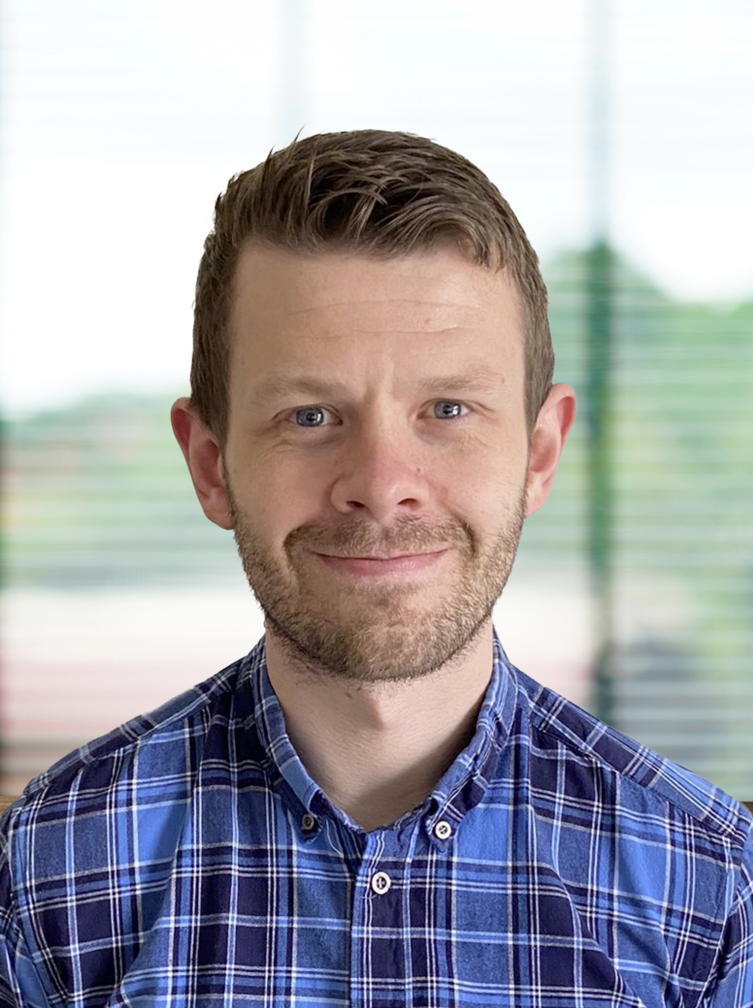 Portrait of a man with short brown hair, light beard, wearing a blue checkered shirt, smiling, in an indoor setting with blurred window blinds in the background.