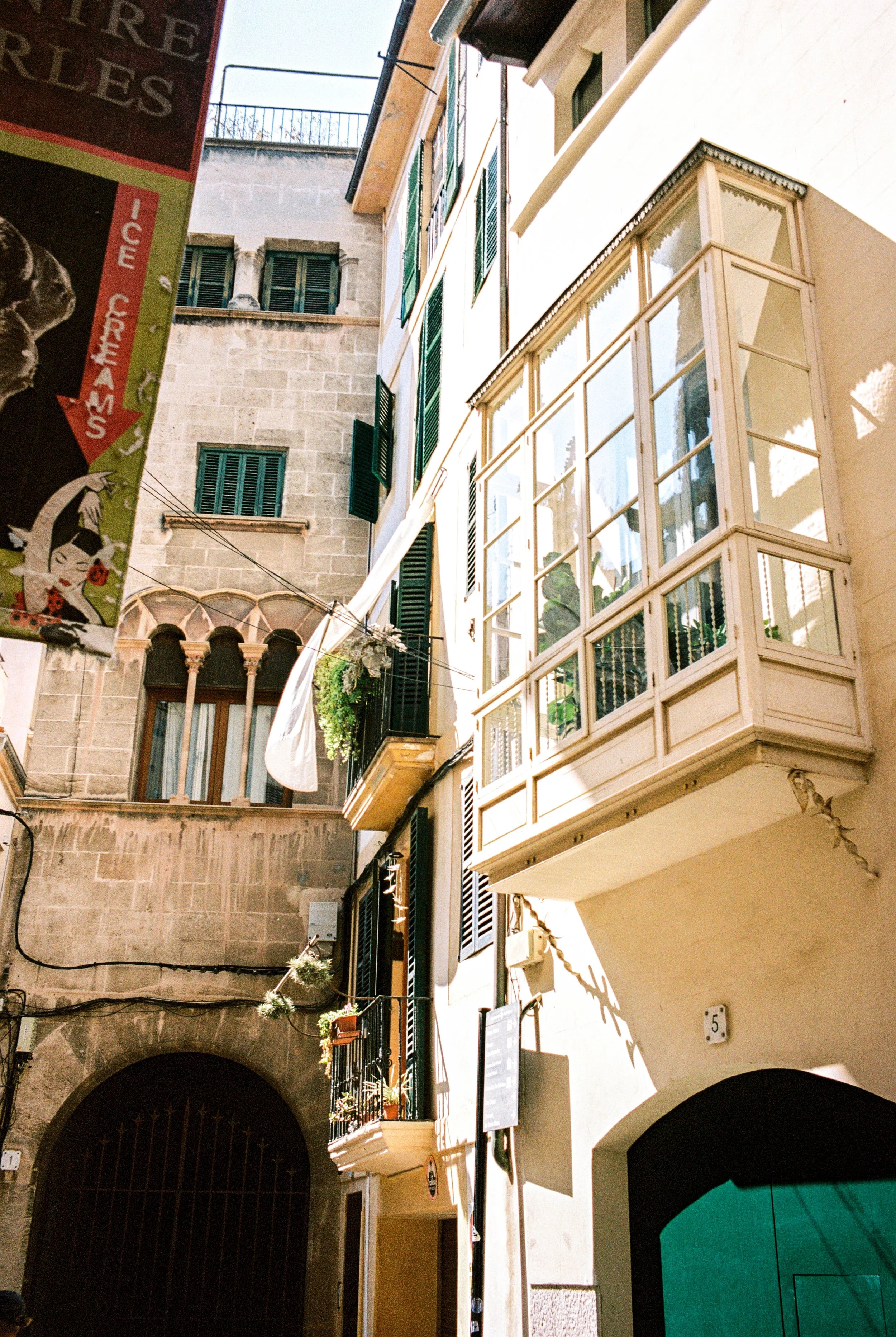 Blick auf enge Straße mit alten Gebäuden, Balkon mit Pflanzen und einer Werbetafel in einer europäischen Stadt.