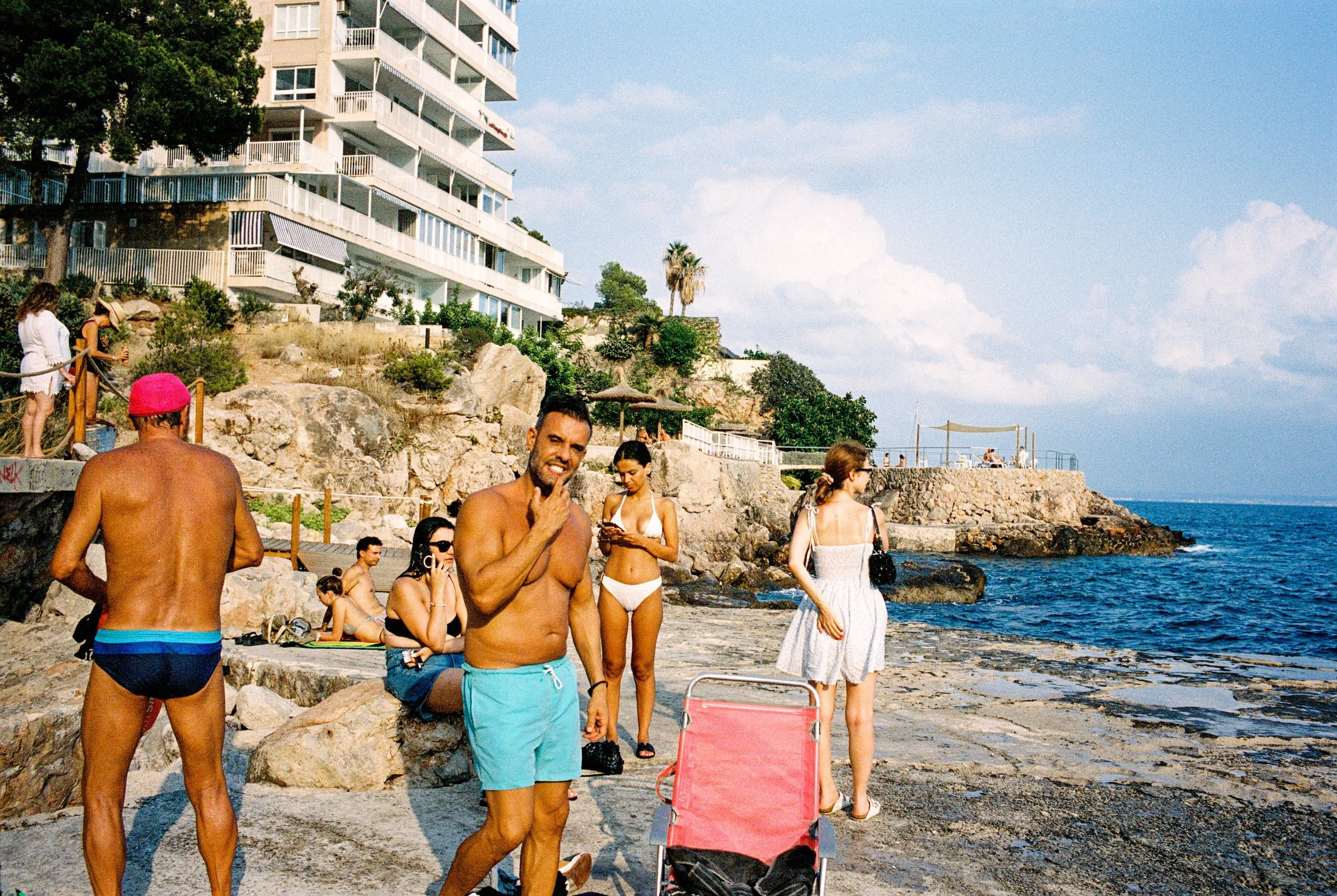 Menschen am Strand, einige sitzen, andere stehen, im Hintergrund ein Hochhaus, Felsen und das Meer, sonniges Wetter.