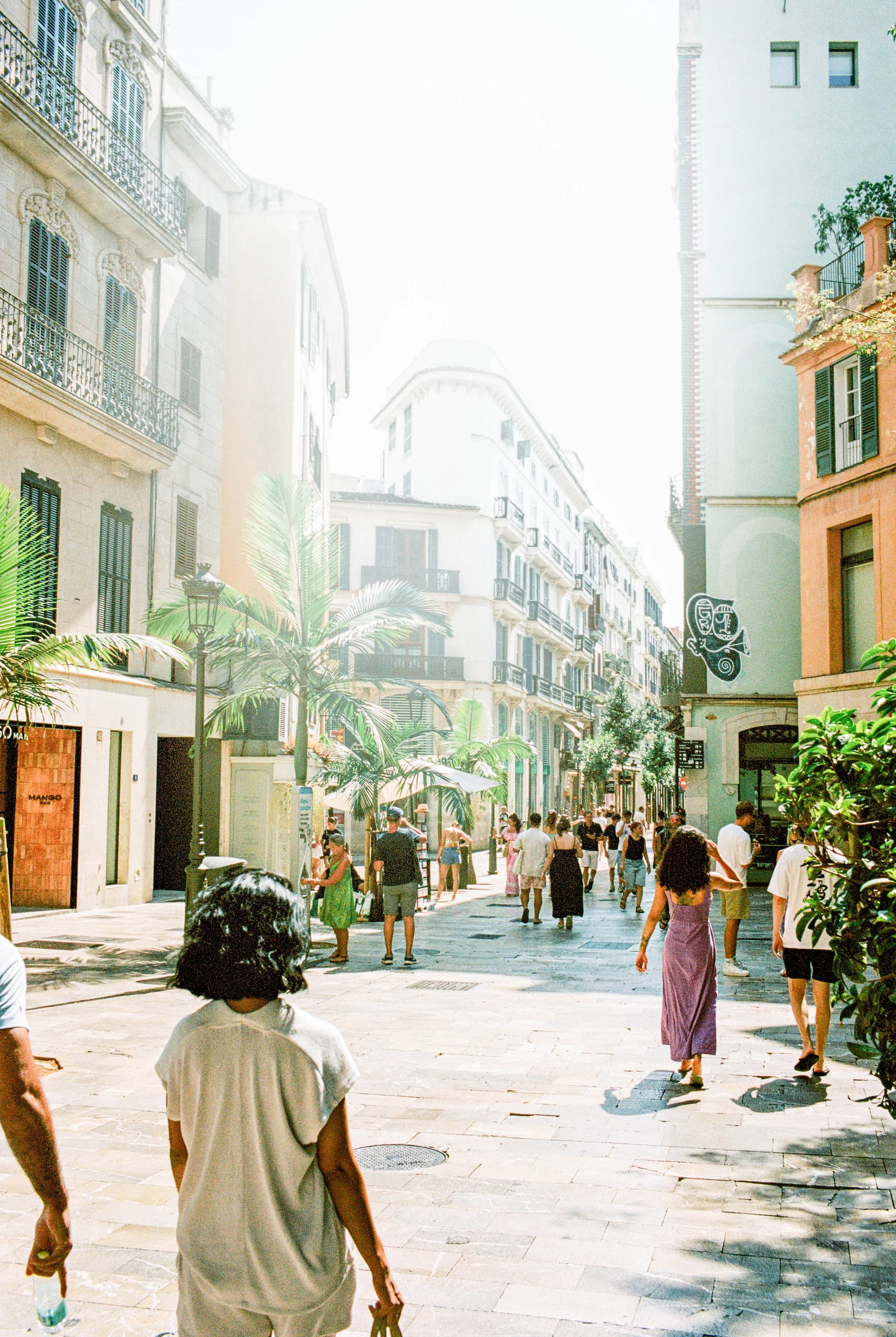 Straße in einer belebten Stadt mit mehreren Passanten, umgeben von mehrstöckigen Gebäuden, Palmen und Straßenschildern, bei sonnigem Wetter.