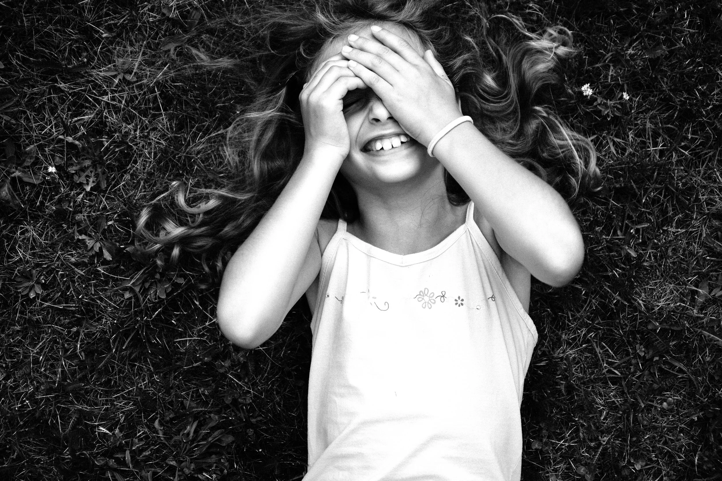 Junge Mädchen liegt lachend auf dem Rasen, macht ein lustiges Gesicht und bedeckt ihre Augen mit den Händen.