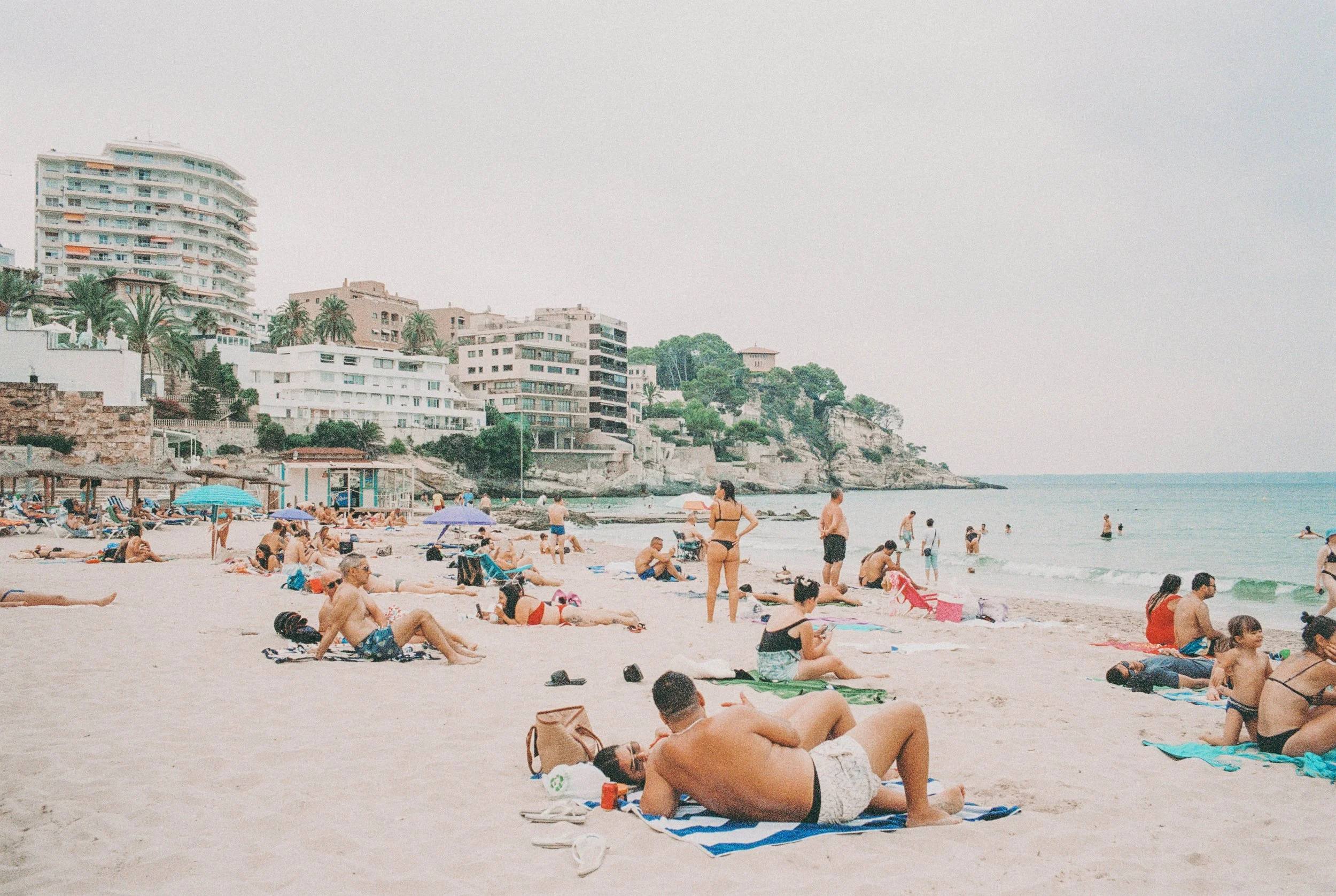 Menschen am Strand mit Gebäuden im Hintergrund, einige liegen auf Handtüchern, andere stehen oder gehen am Wasser.