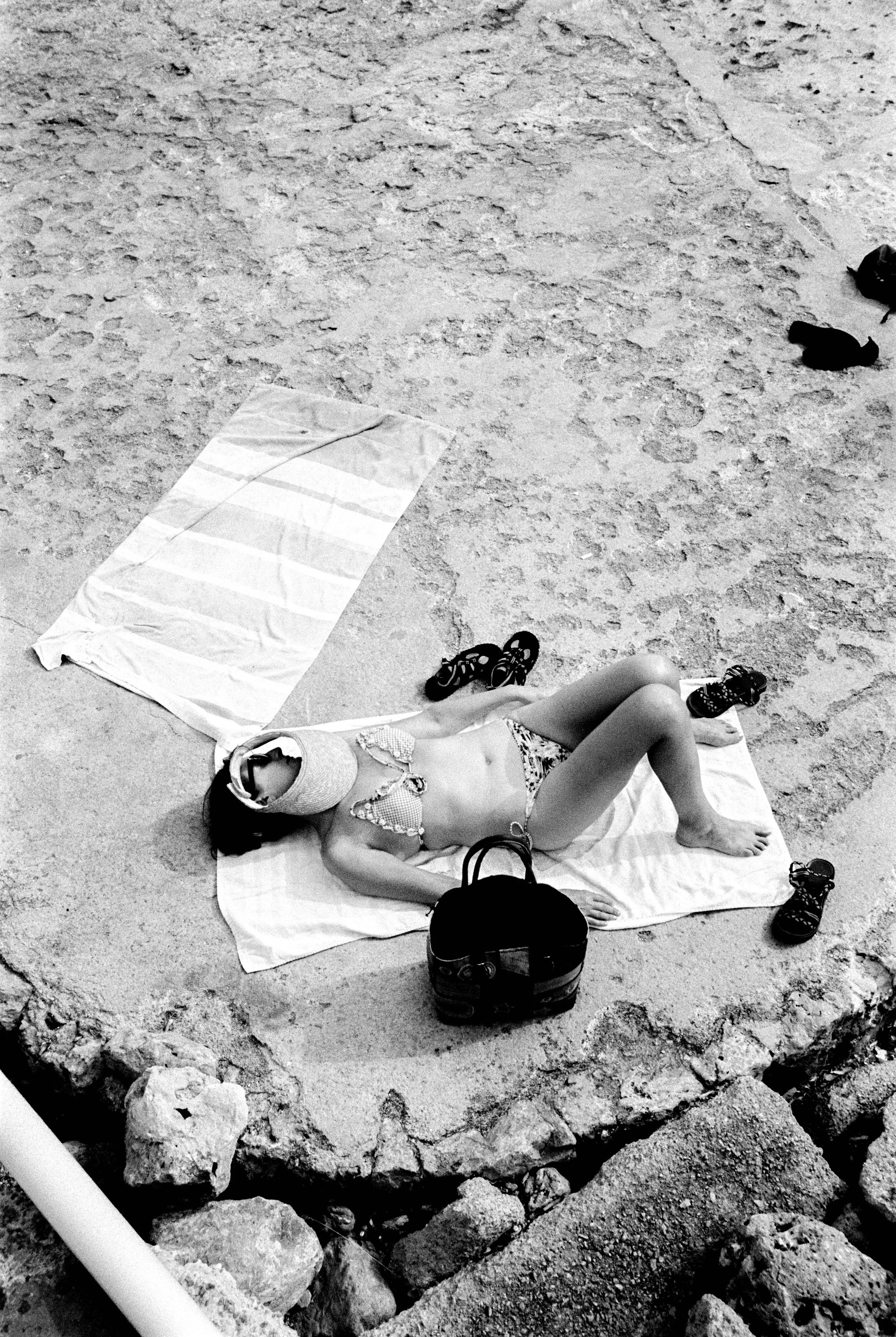 Frau liegt sonntags auf der Strandmatte, trägt Sonnenbrille und Bikini, umgeben von Schuhe und einer Tasche, im Sand. Das Bild ist schwarz-weiß.