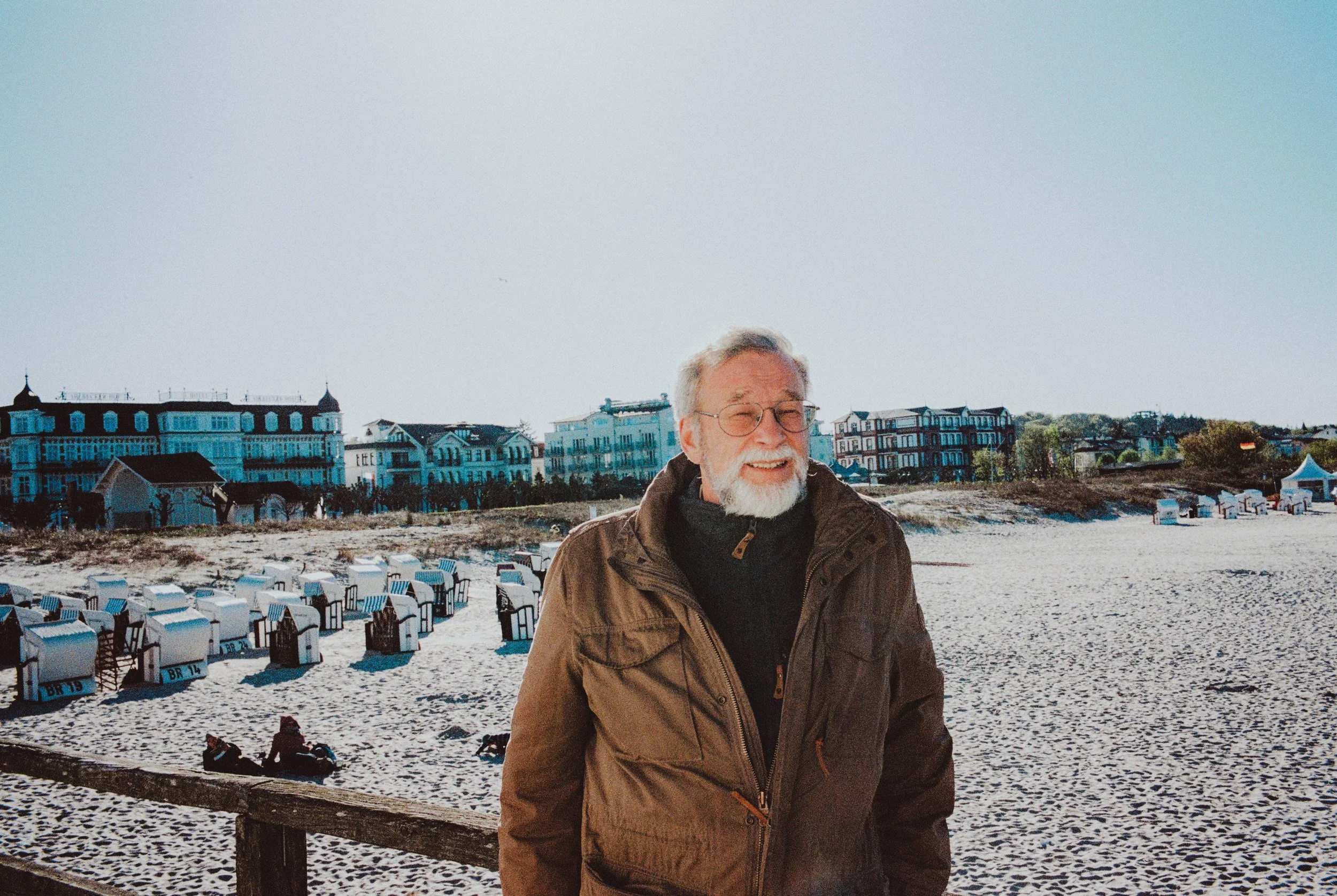 Ein älterer Mann mit Brille und grauem Bart steht am Strand mit Strandkörben, im Hintergrund bunte Gebäude und ein Himmel, der auf einen sonnigen Tag hinweist.