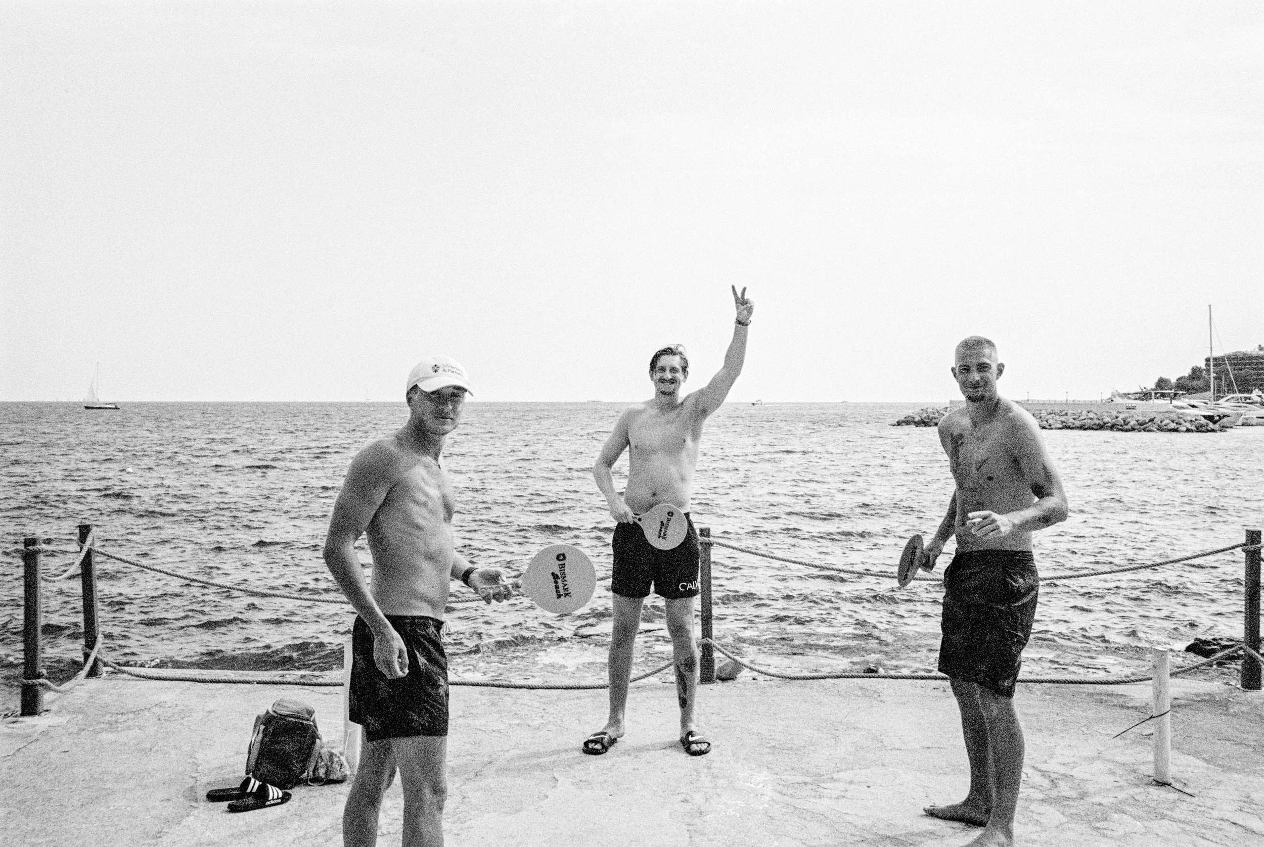 Drei Männer in Badekleidung mit Paddeln am Wasser, einer macht ein Peace-Zeichen, am Strand in der Nähe eines Meeres