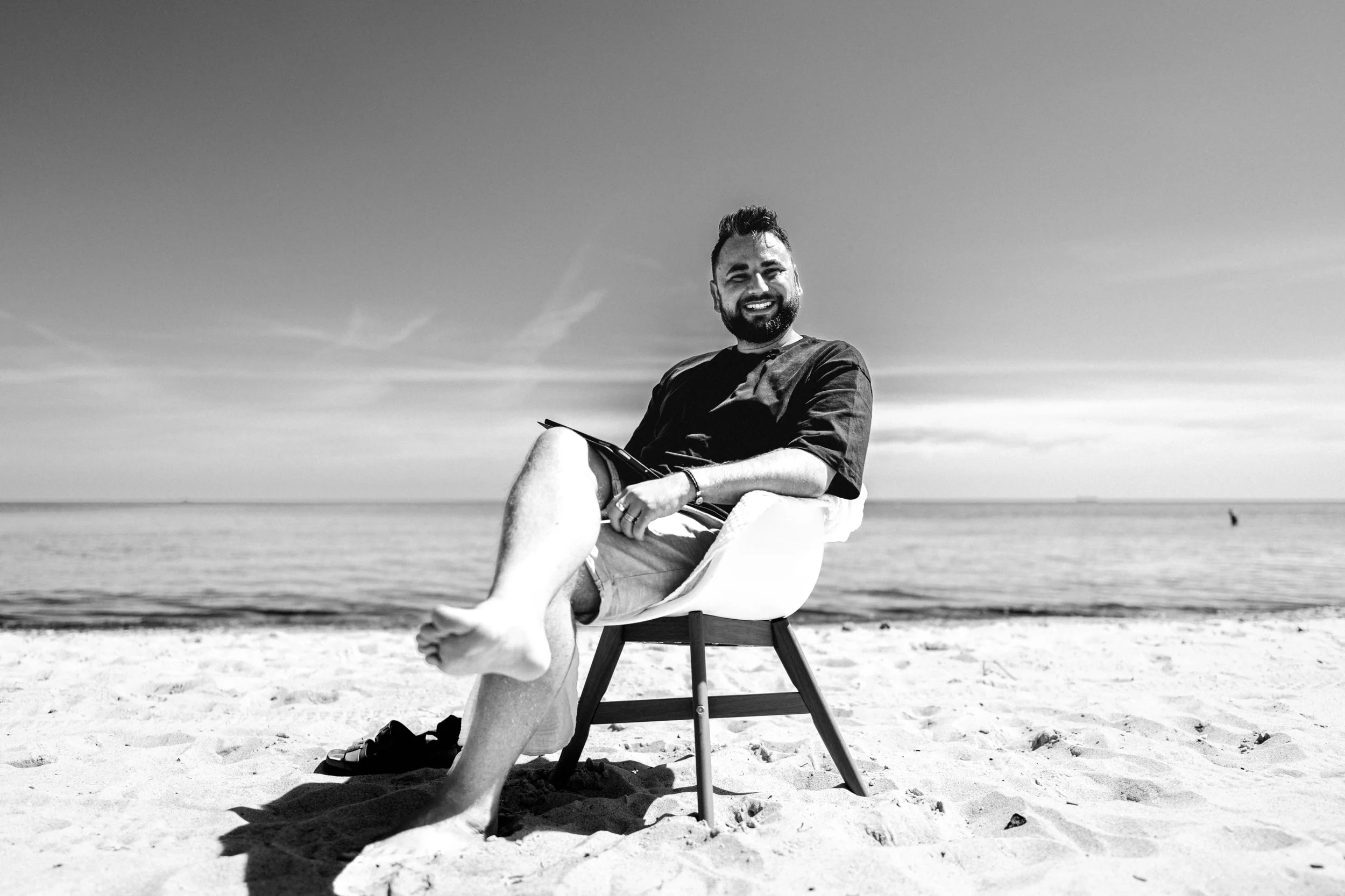 Mann sitzt entspannt in einem Stuhl am Strand, im Hintergrund das Meer, in Schwarz-Weiß.