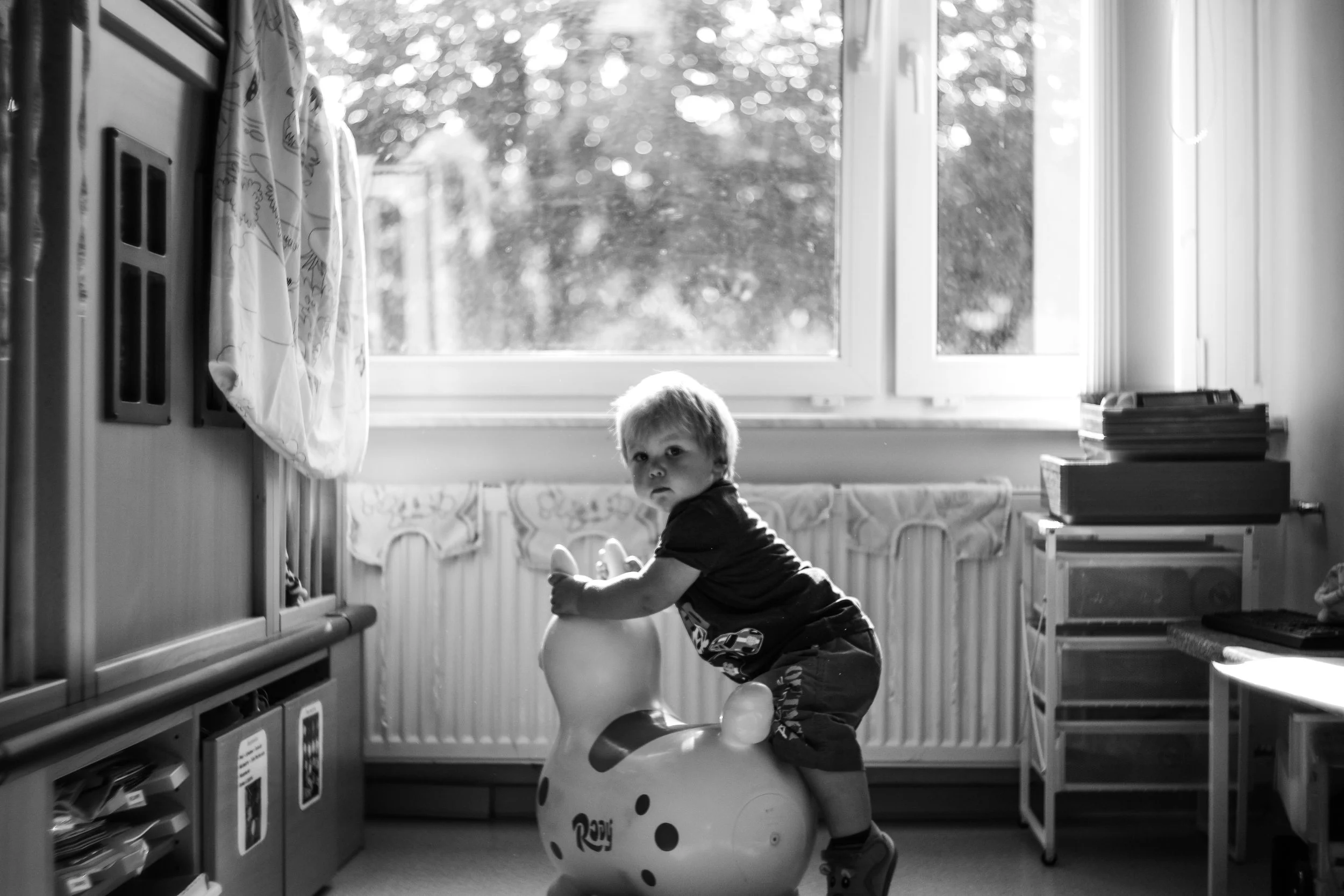 Ein Kind sitzt auf einem plüschigen Spielzeug im Wohnzimmer, blickt zur Kamera, mit einem Fenster im Hintergrund, das Licht ins Zimmer lässt, in schwarzer und weißer Fotografie.