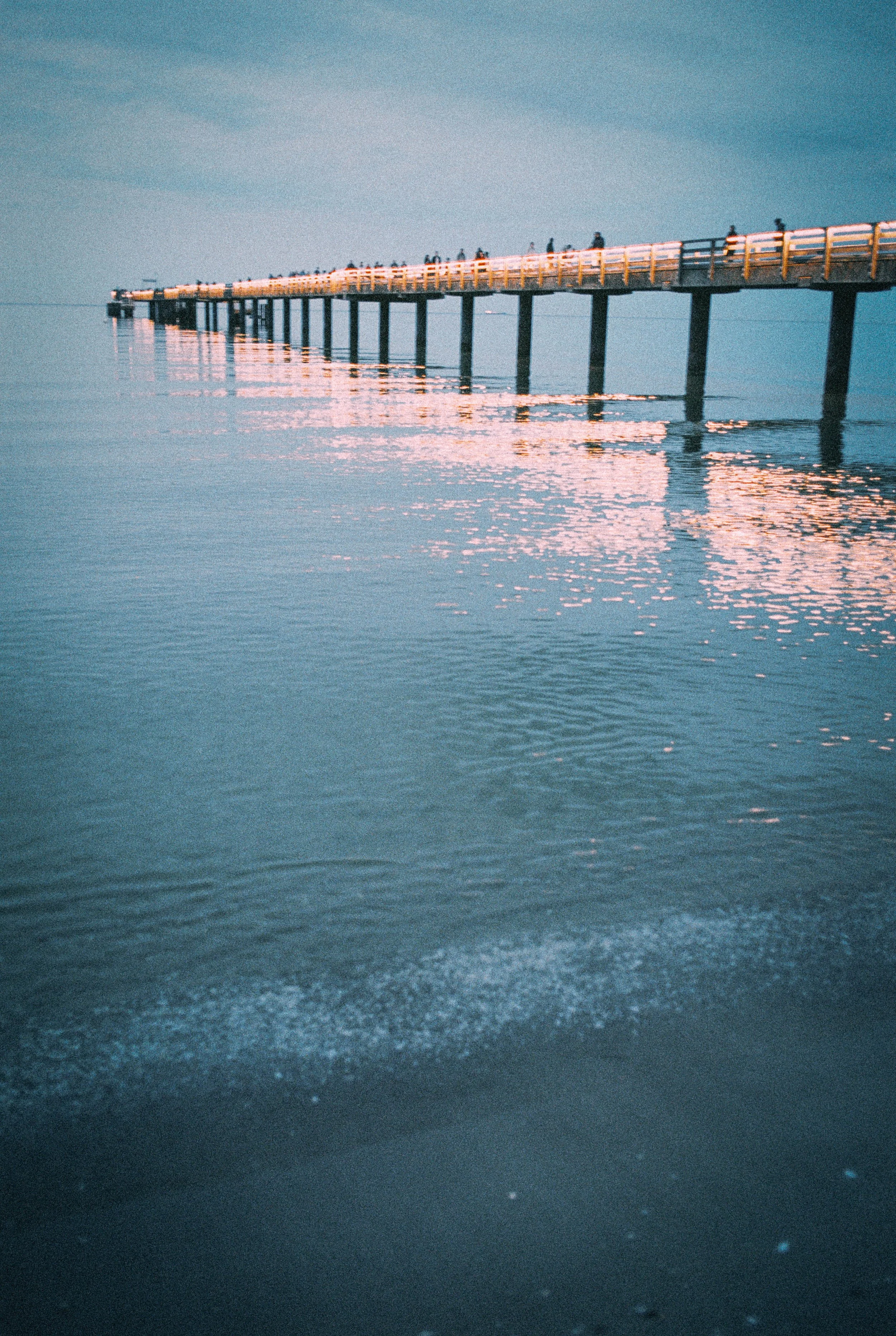 Ein langer Steg, der ins Meer führt, bei Sonnenuntergang, mit Menschen, die entlang gehen, Reflexionen im Wasser.