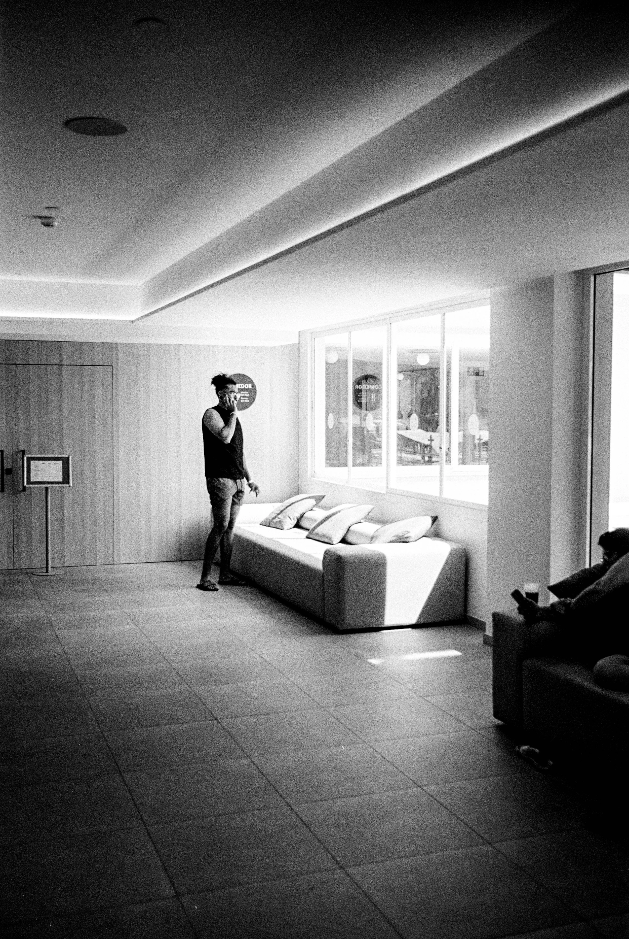 Schwarz-Weiß-Foto eines Wartebereichs in einem Gebäude mit zwei Personen, die einen Stuhl benutzen, einer Person steht near das Fenster, die andere sitzt an einem Sofa und liest auf einem Handy.