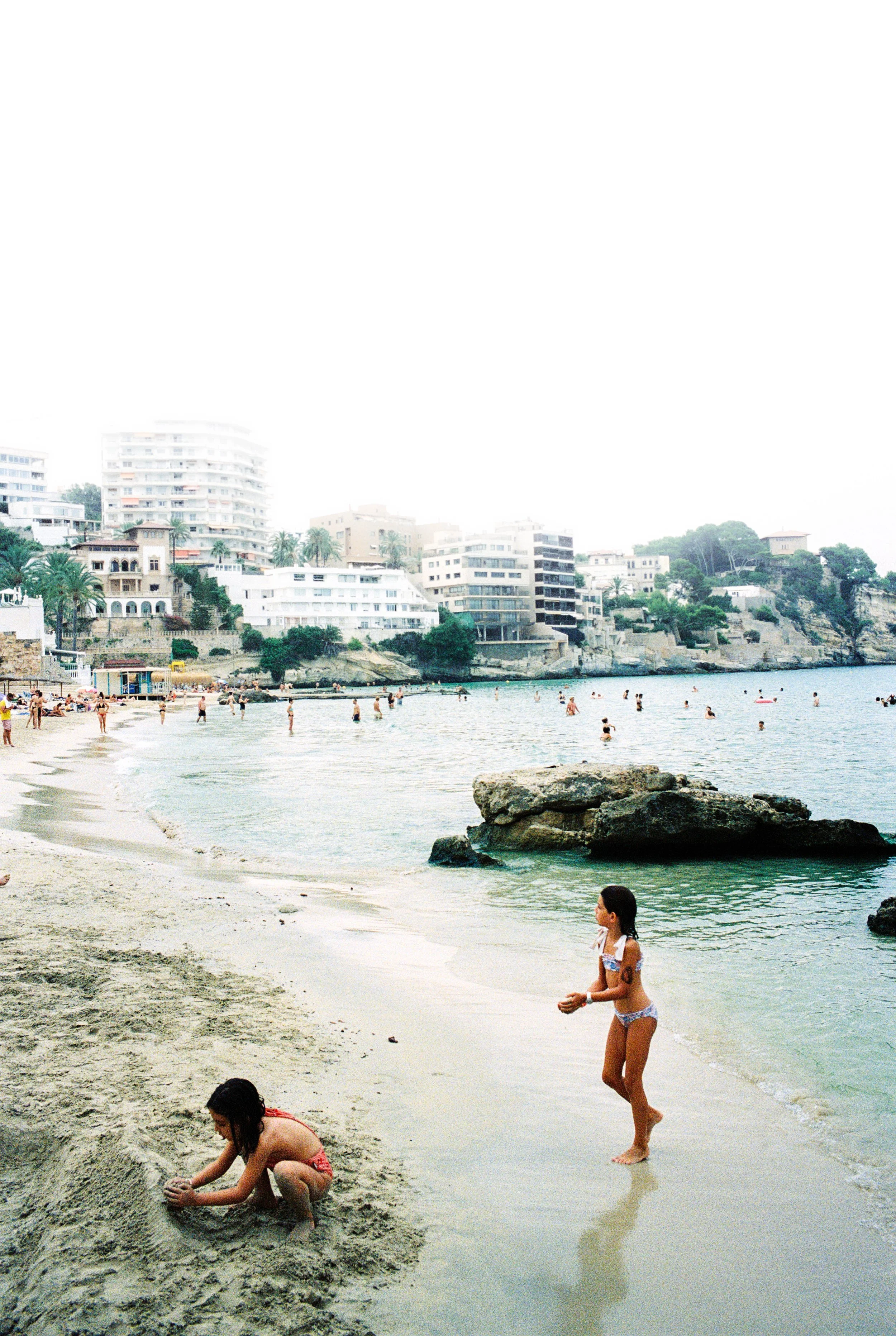 Kinder spielen am Strand mit Sand und Wasser, im Hintergrund sind weiße Gebäude und menschenleere Stadtflächen.