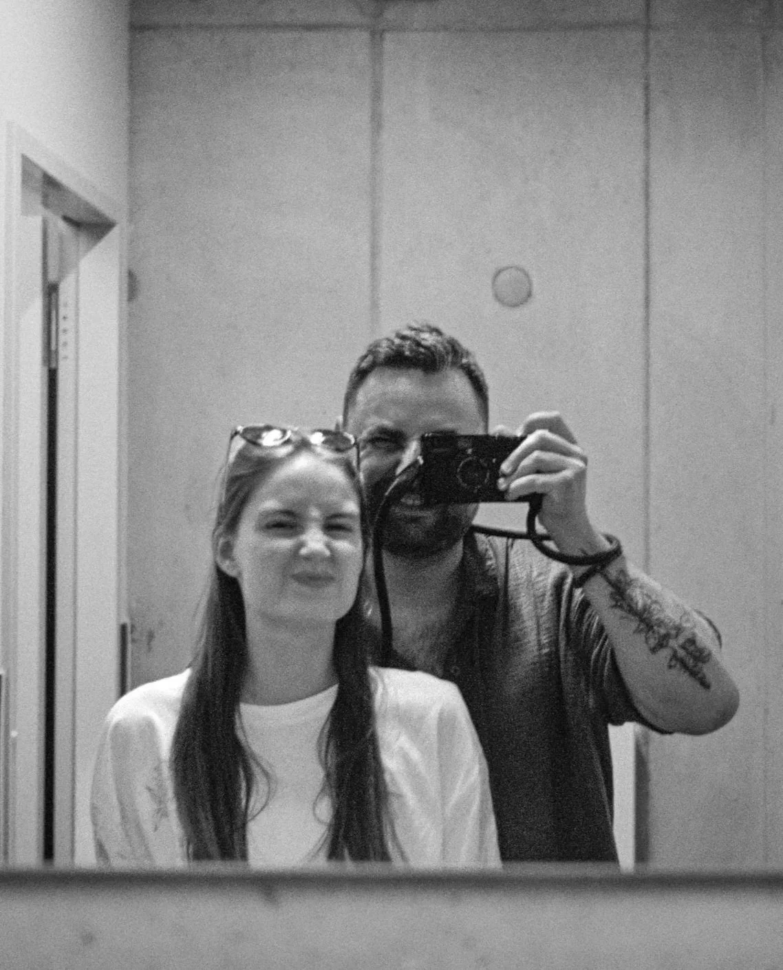 Ein Mann und eine Frau nehmen ein Selfie vor einem Spiegel auf, schwarz-weiß, in einem Raum mit glatten Wänden.