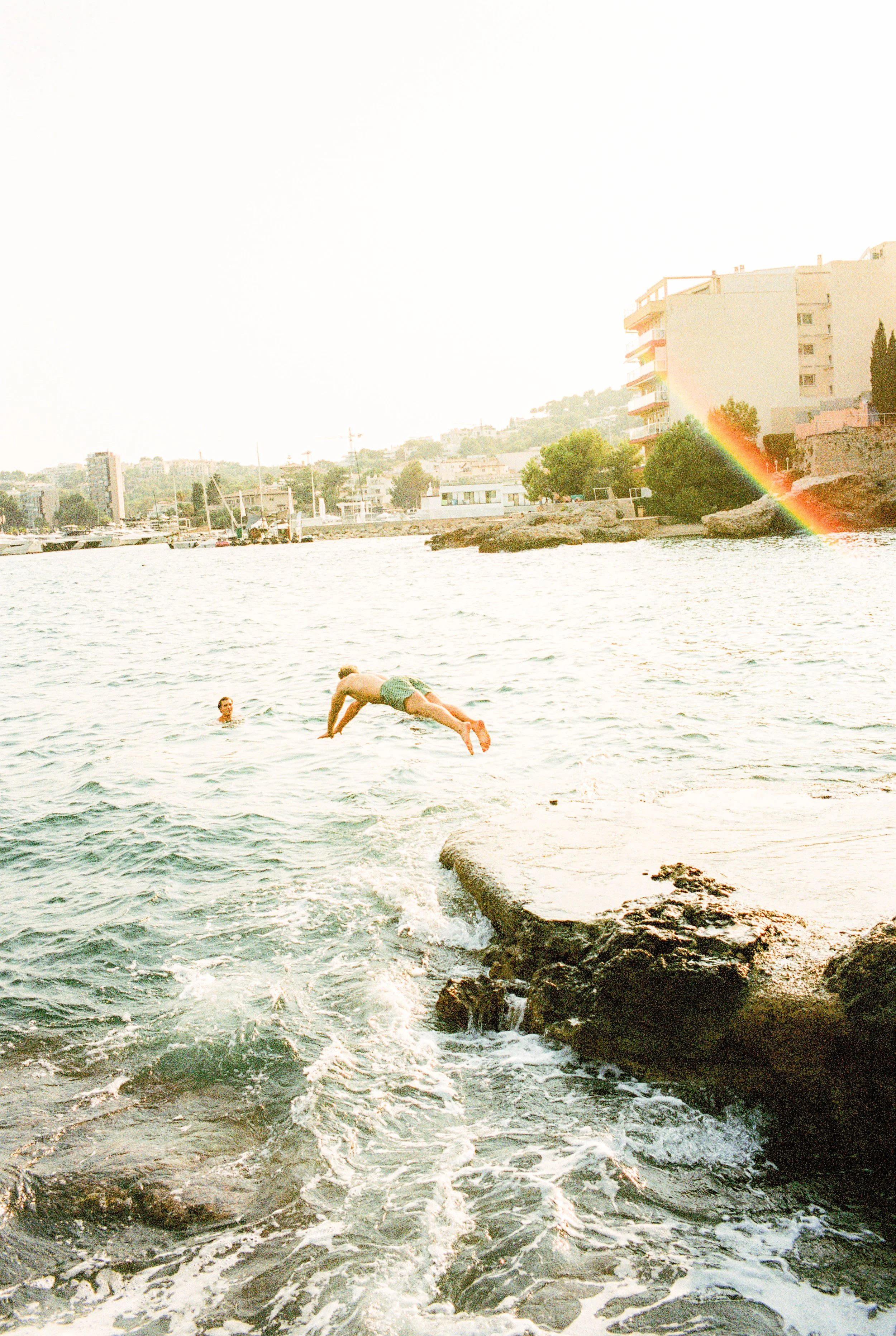 Person springt vom Felsen ins Wasser, andere Person schwimmt im Wasser, Stadt und Regenbogen im Hintergrund.