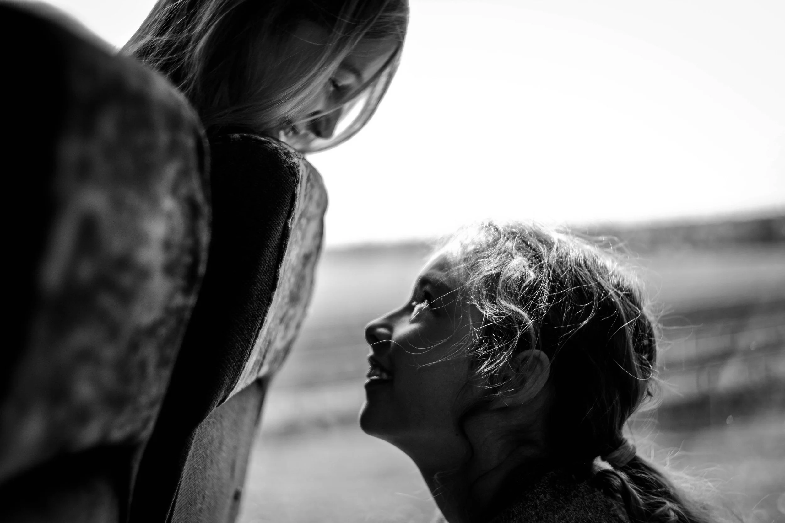 Zwei junge Mädchen schauen sich lachend an, eines schaut über eine Sitzlehne hinweg. Das Bild ist schwarz-weiß und zeigt eine emotionale Szene draußen bei Tageslicht.