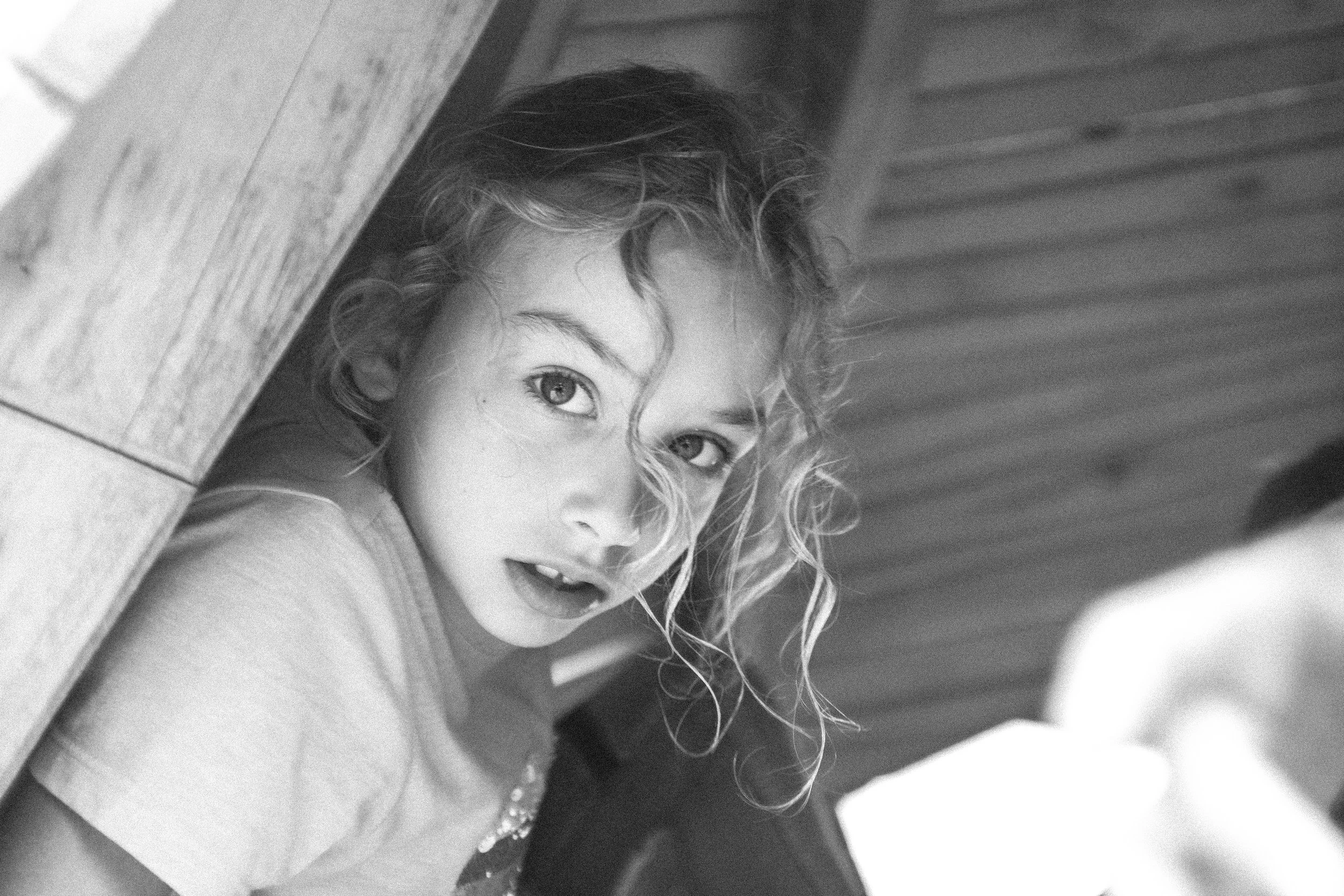 Schwarz-Weiß-Foto eines Mädchens mit lockigem Haar, das seitlich hinter einer Holztafel hervorschaut.