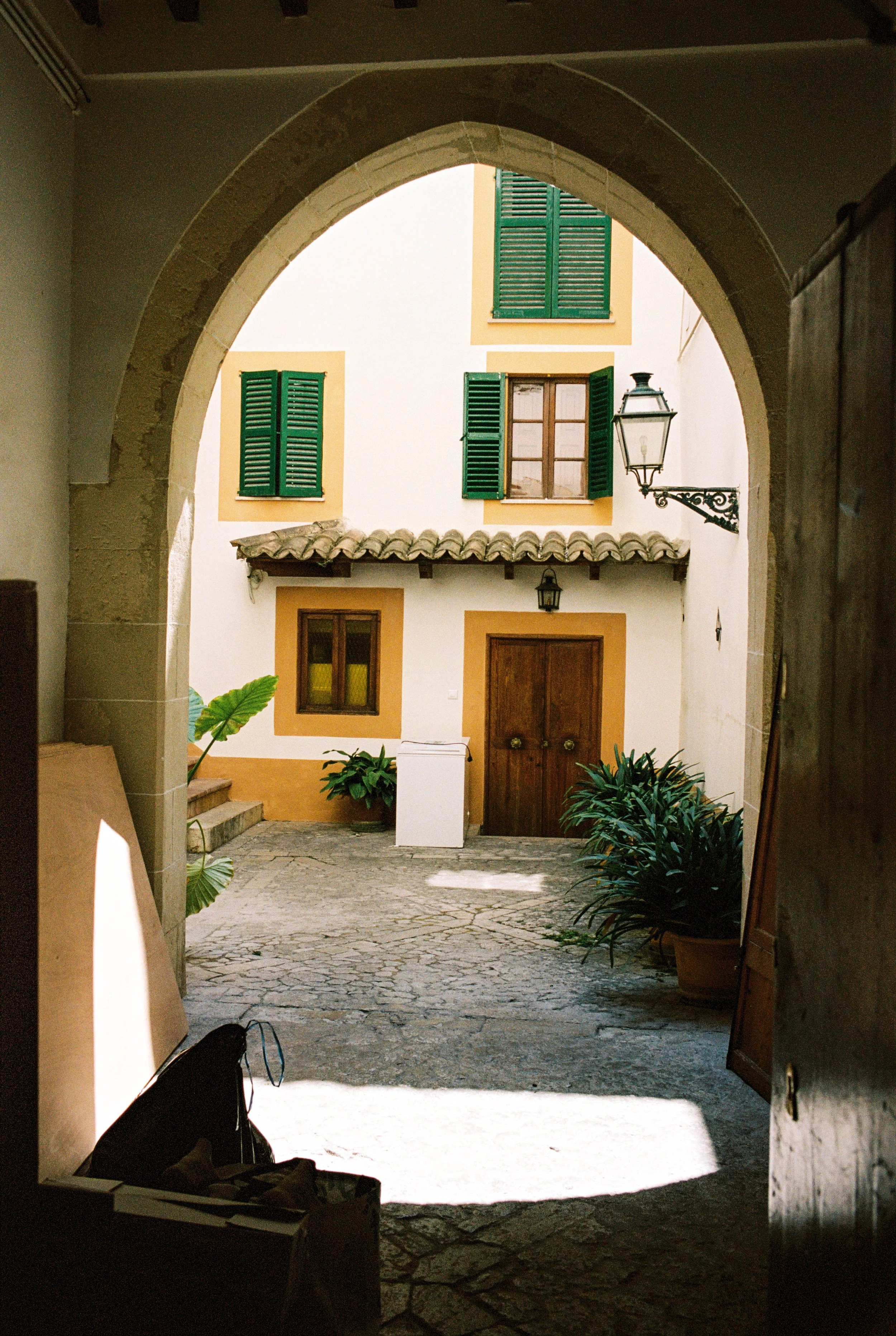 Blick durch einen Torbogen auf einen Innenhof mit weißen Wänden, grünen Fensterläden, einer Holzhaustrat und Pflanzen.