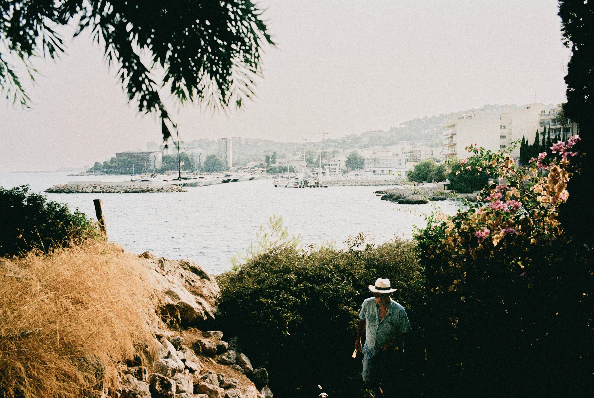 Ein Mann mit weißem Hut und Sonnenbrille, in einem blauen Hemd, läuft durch ein bewaldetes Gebiet mit Blüten und Sträuchern in der Nähe eines Hafens mit Booten und Gebäuden im Hintergrund.