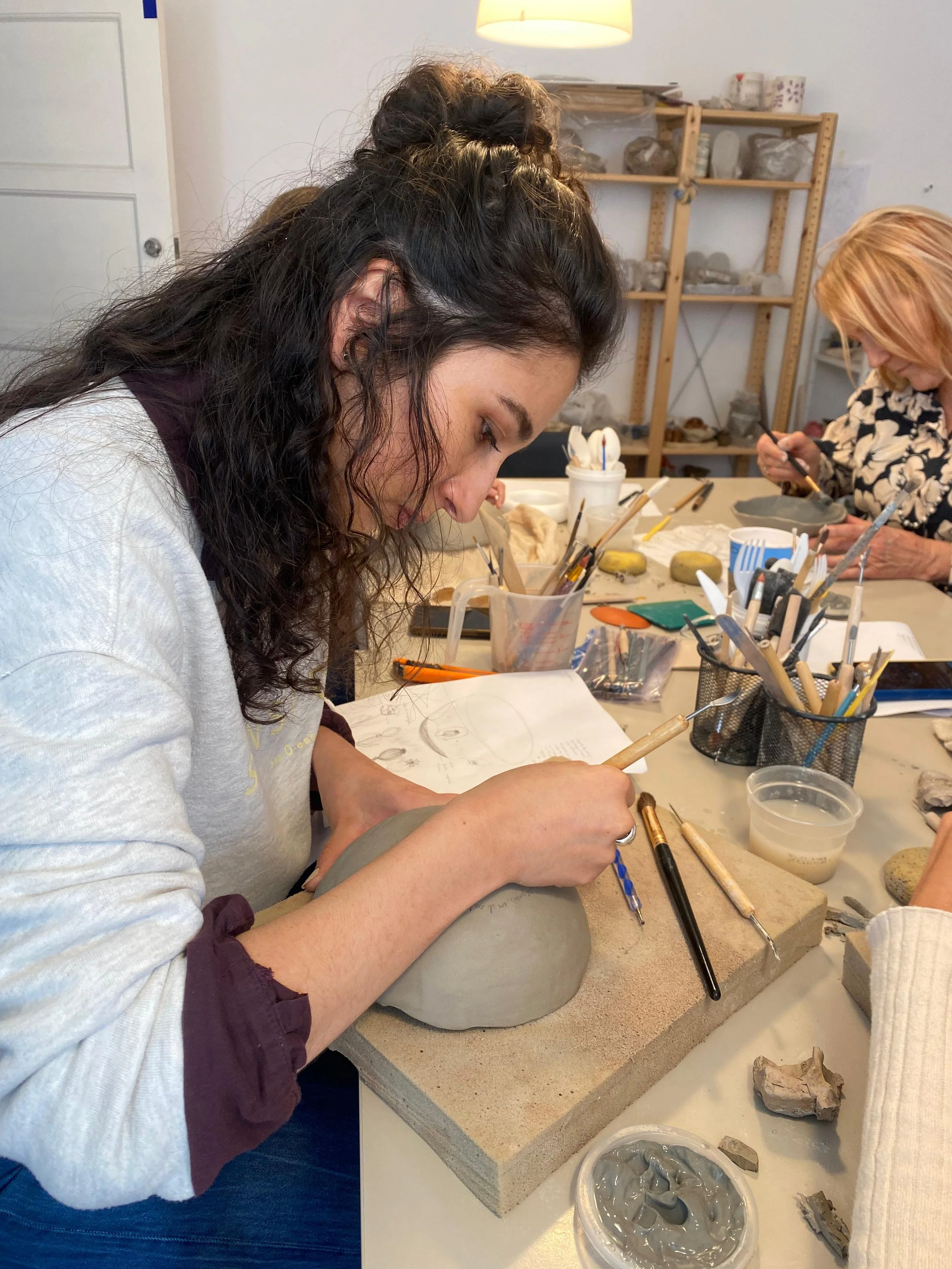 Mujer joven trabajando en cerámica en un taller, con herramientas y piezas de arcilla en la mesa.