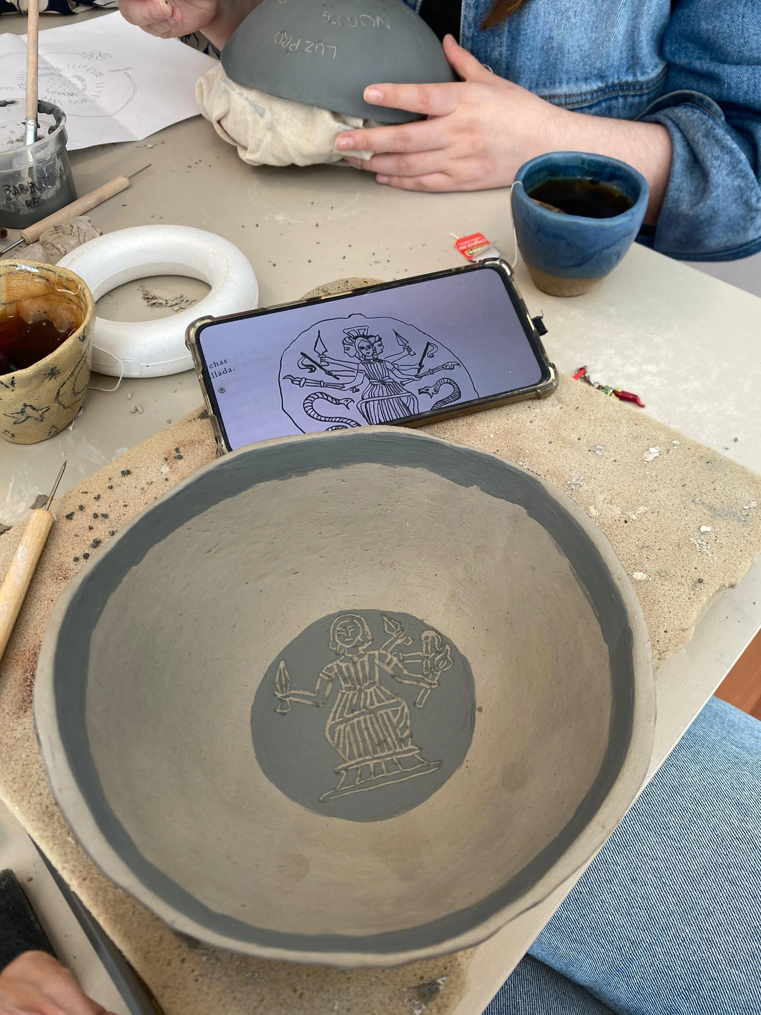 Vasos de cerámica con decoraciones, herramientas de cerámica, una persona sosteniendo una pieza de cerámica y un teléfono móvil con un dibujo de una figura mitológica en la pantalla, en una mesa de trabajo de cerámica.
