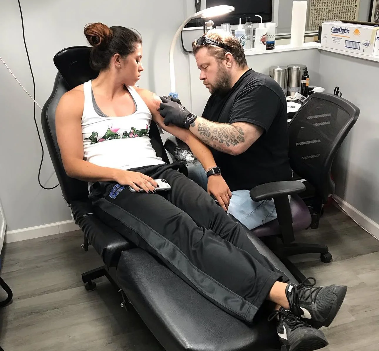 A woman receiving a tattoo on her upper arm from a male tattoo artist in a tattoo studio. The woman is sitting on a black chair, holding a phone, and the artist is wearing black gloves and working with tattoo equipment.