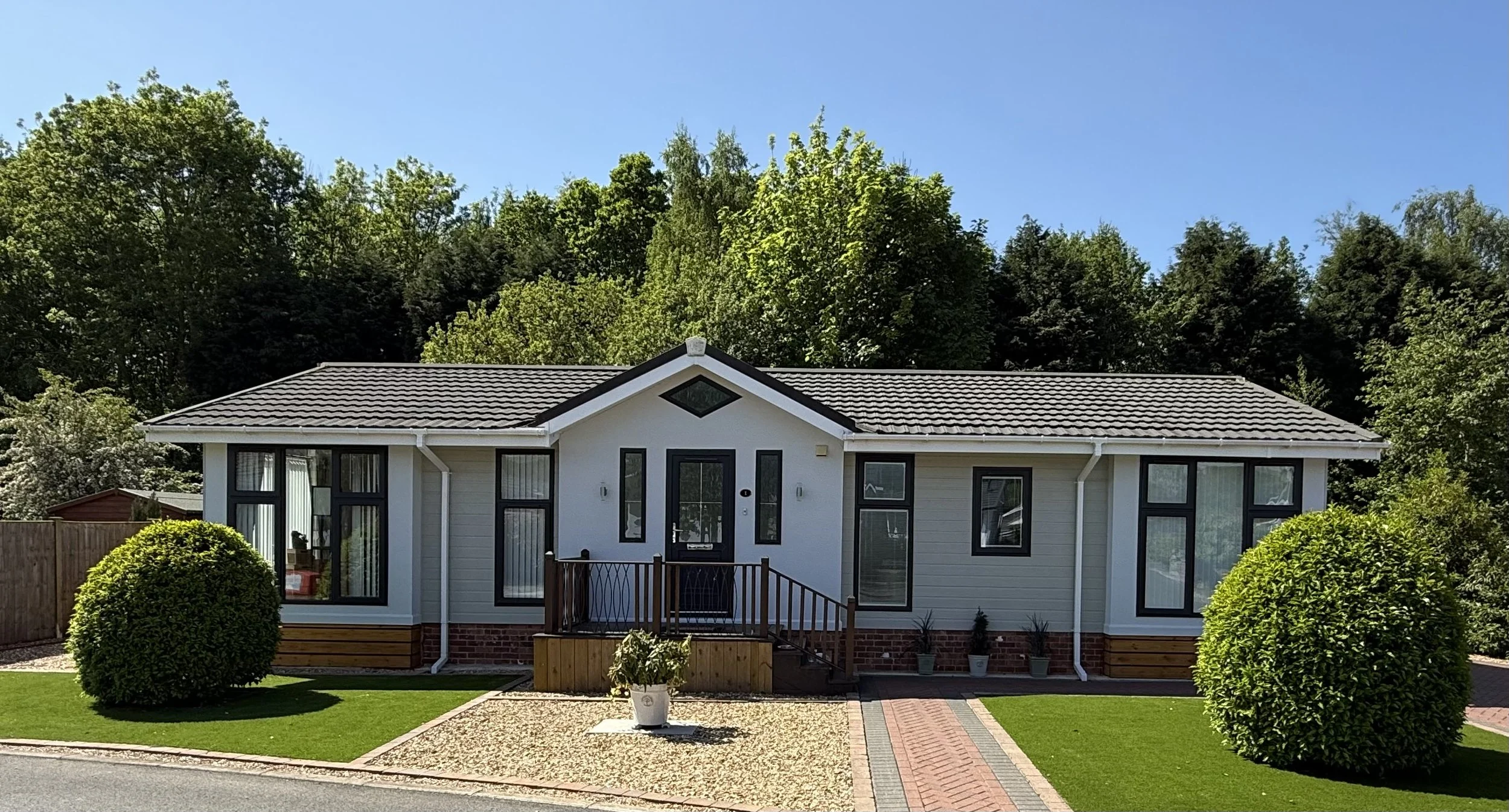 A modern single-story house with white siding, black window frames, and a gray tiled roof. The house has a small front porch with wooden steps and railings. There is a well-maintained lawn with two large round bushes on either side of a gravel pathway and a brick walkway. Green trees and a clear blue sky are in the background.