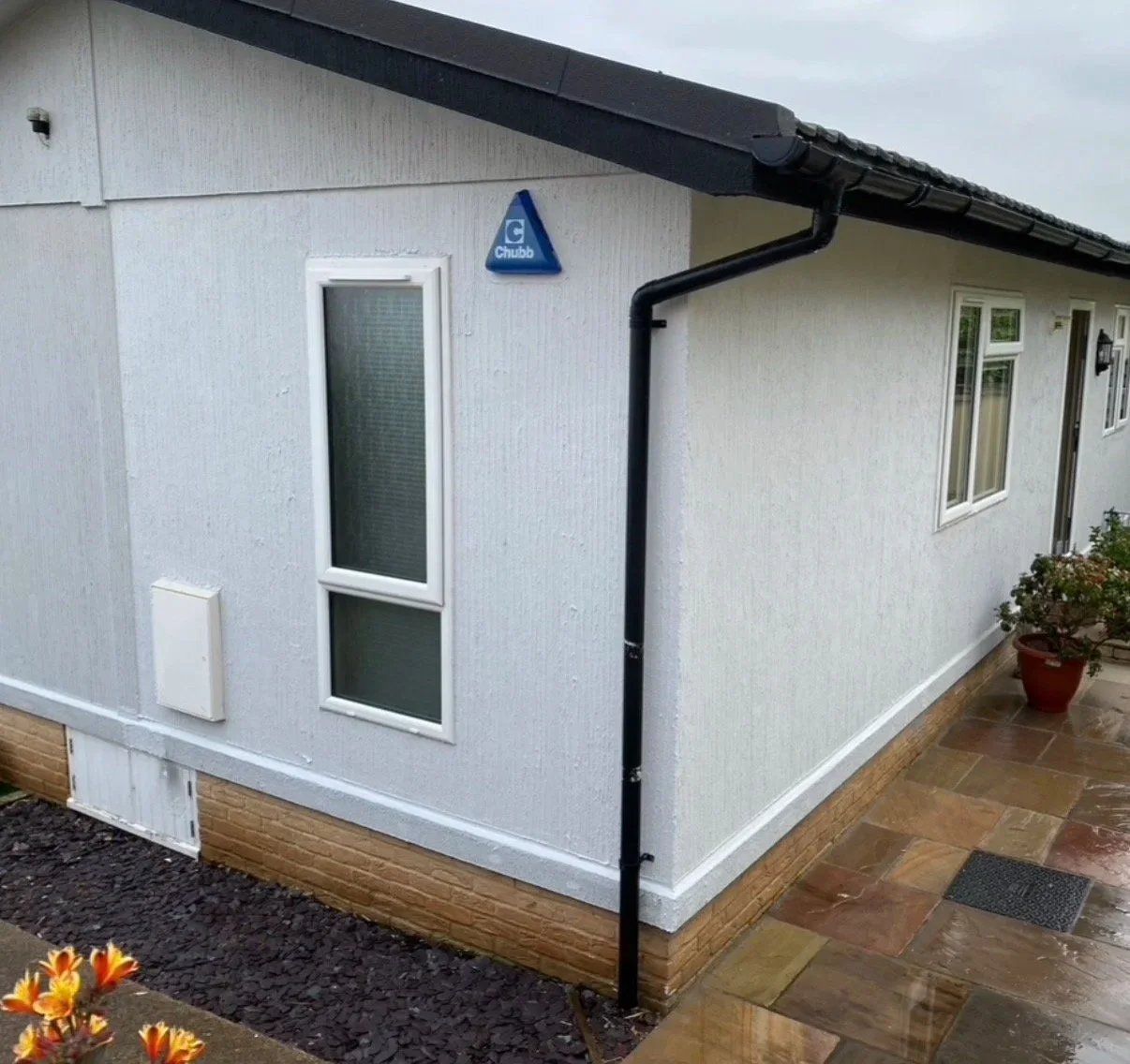 Exterior corner of a house with white textured walls, black gutter system, and small rectangular windows. Potted plant on a paved walkway, and a blue triangular Chubb security sign on the wall.