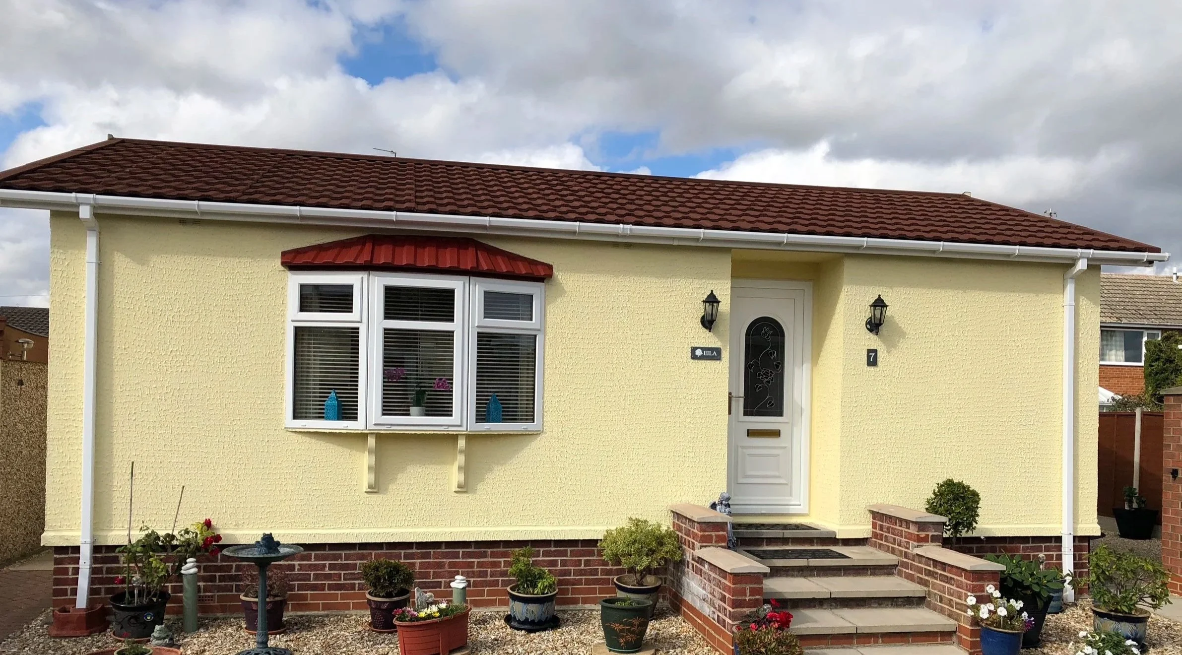 A yellow house with a red-tiled roof and a white front door, side wall lanterns, a bay window with red awning, and potted plants outside on a gravel yard.