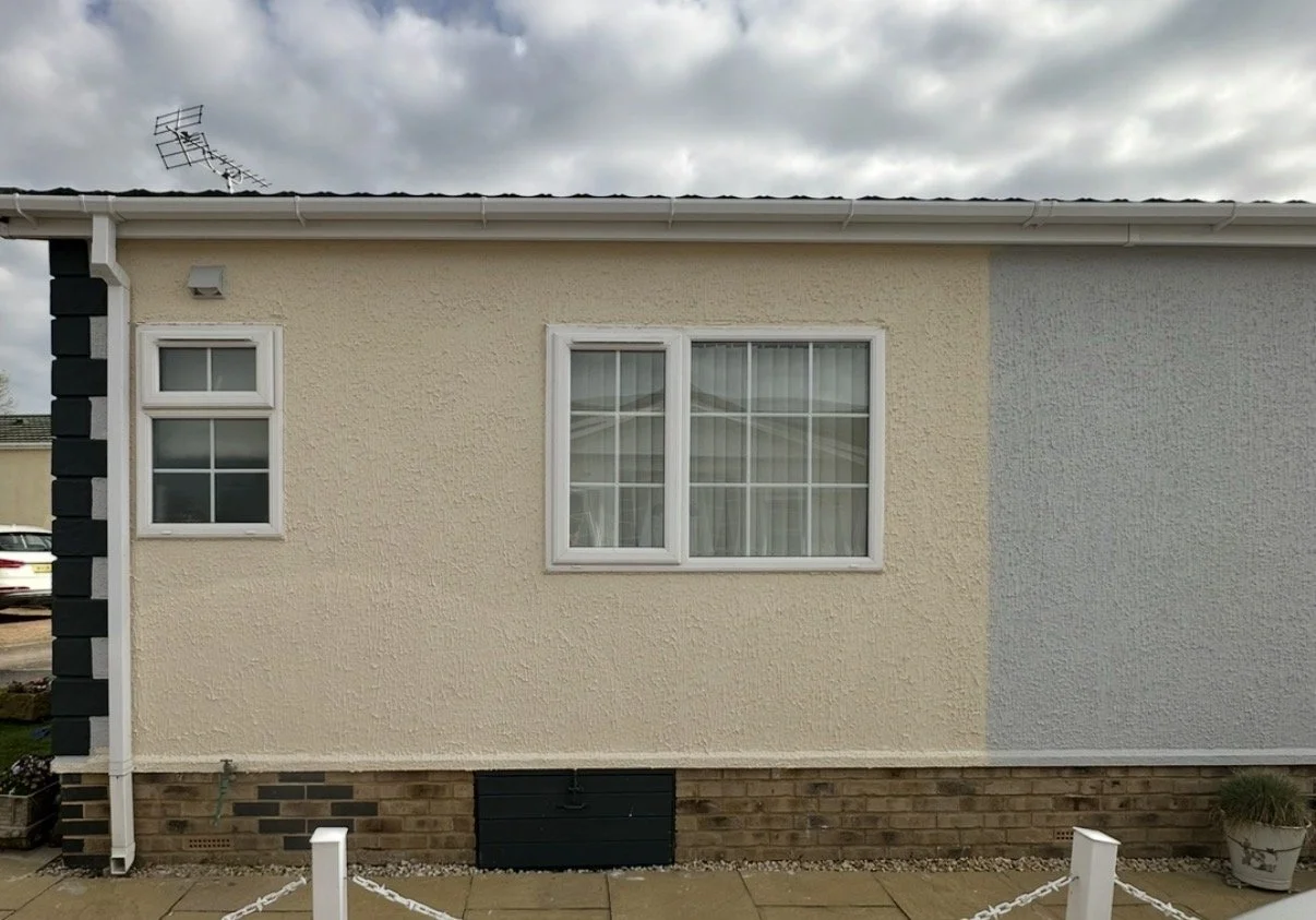 A beige and gray textured house exterior with three windows, one small and two large, with white frames. The sky above is cloudy.