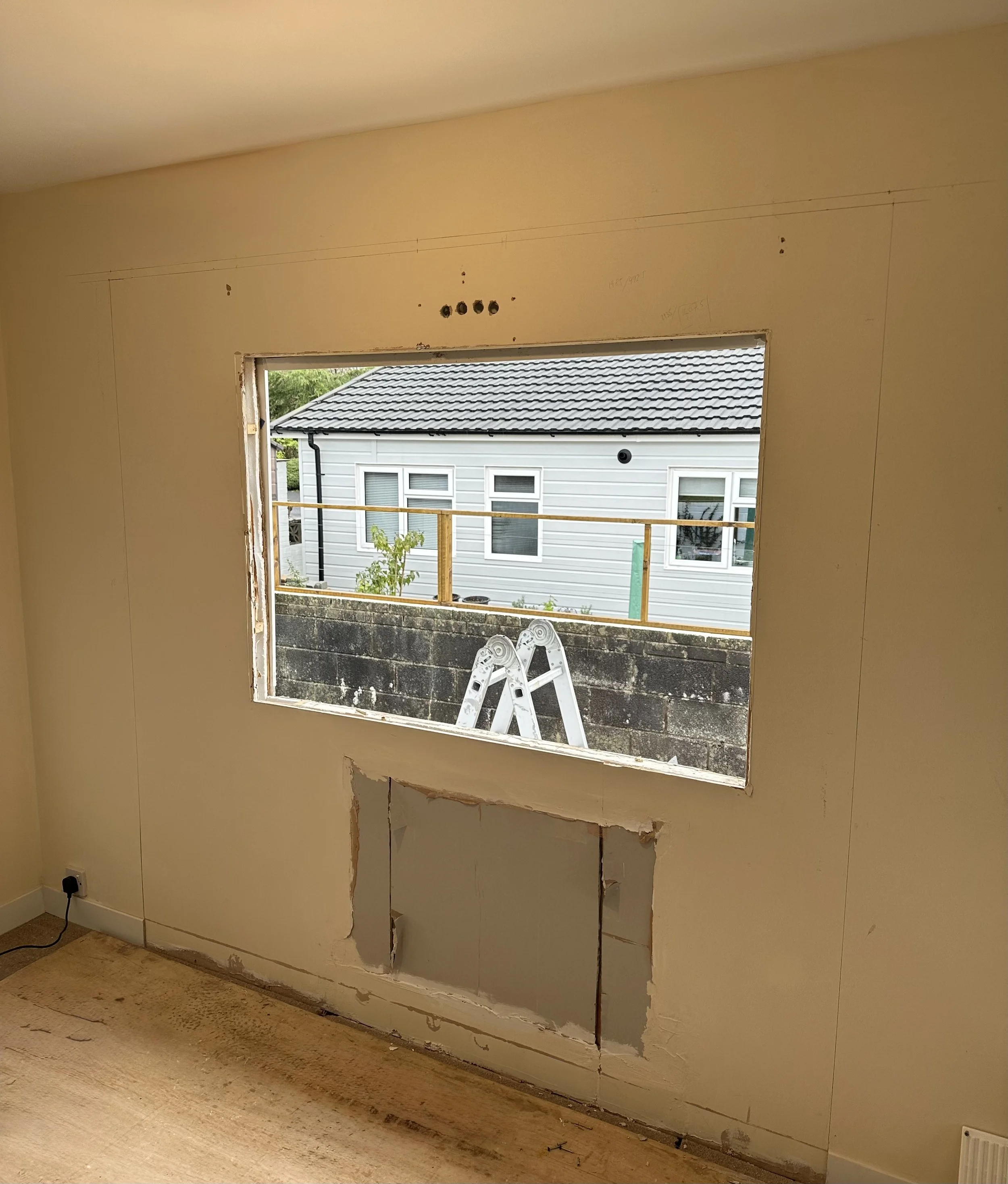 An interior room with a large rectangular window opening under construction, showing a neighboring house with white siding and multiple windows, a ladder outside, and a small tree.