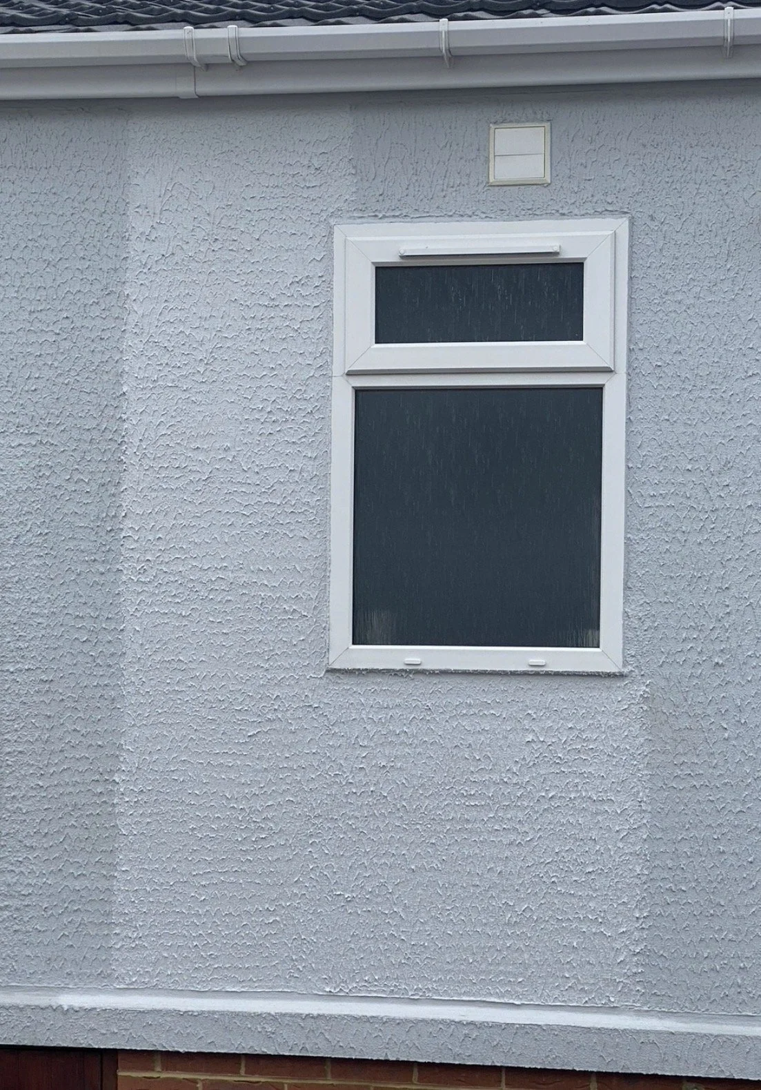 Exterior wall with a textured white stucco finish, a rectangular window with a white frame and a smaller rectangular section above, and a gutter along the roof edge.