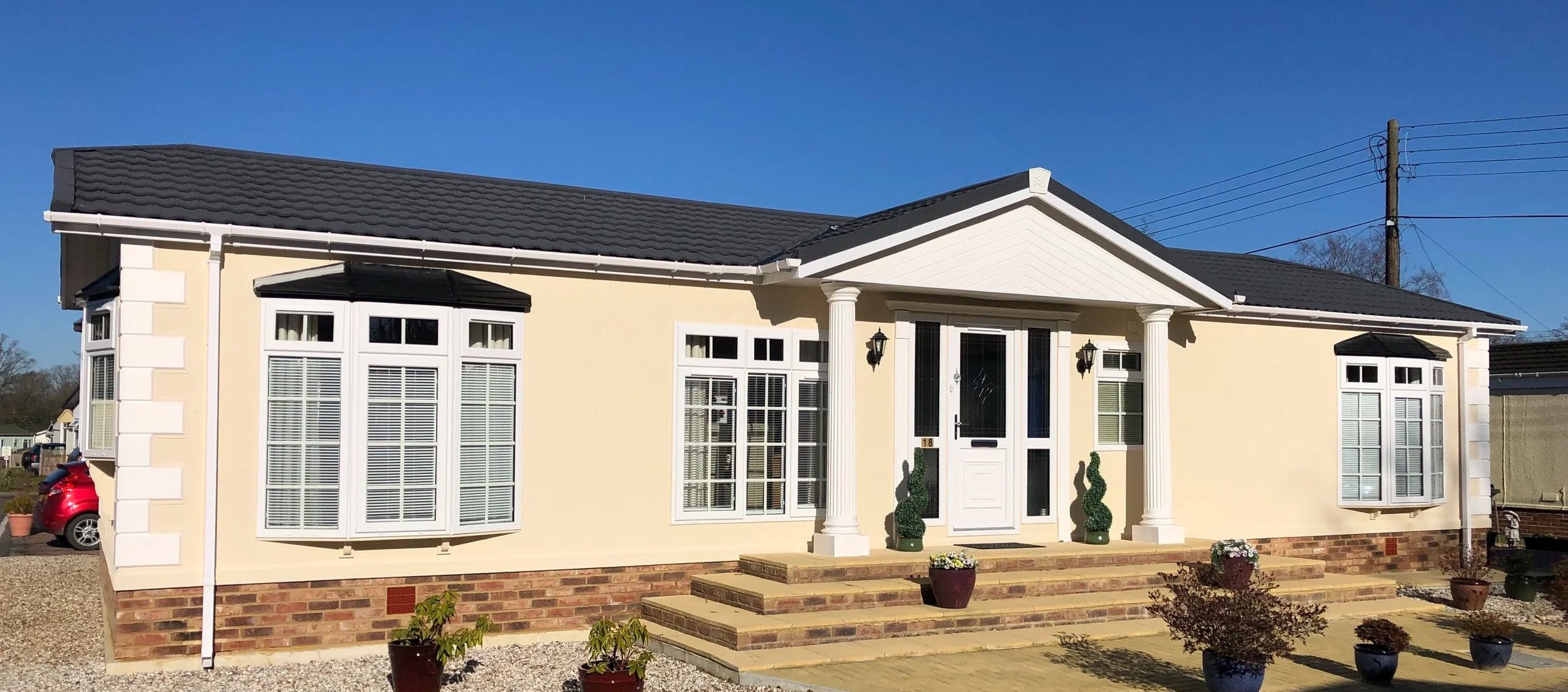 A single-story suburban house with a yellow exterior, white columns, and brick steps leading to the front door. Multiple windows with white shutters and potted plants in the front yard. Clear blue sky in the background.