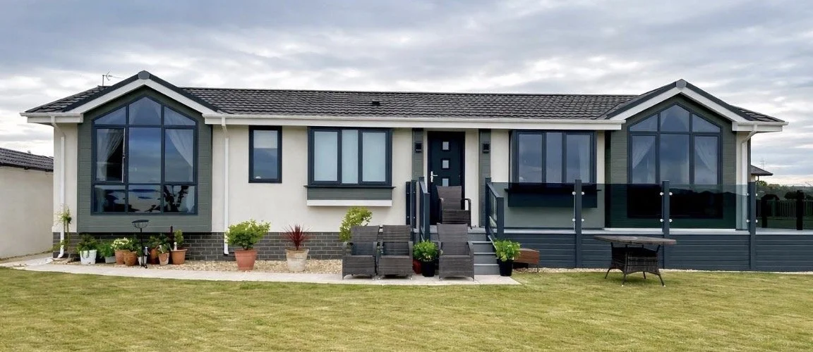 Modern suburban house with large glass windows, a grey exterior, a raised deck with black railings, and a well-maintained lawn.