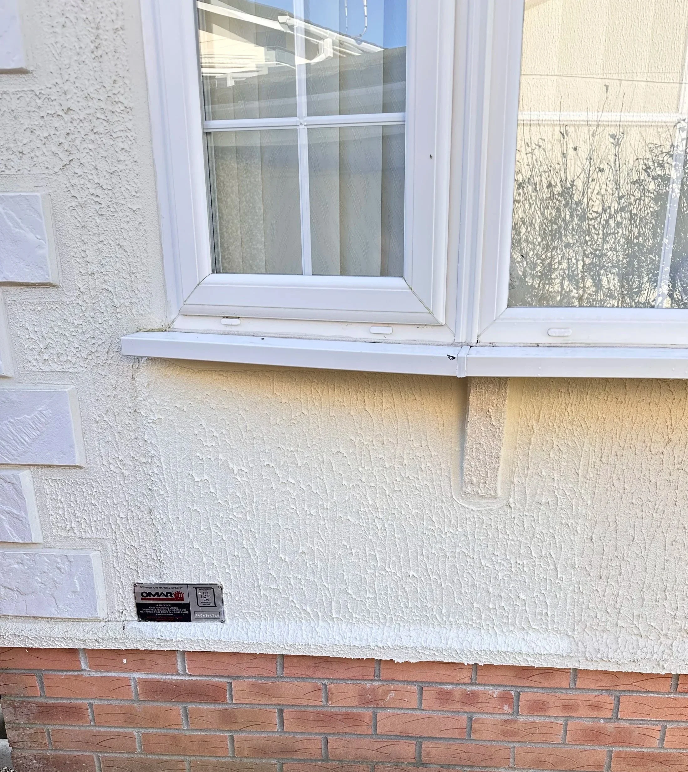 Close-up of a house wall showing a window with white trim, textured cream-colored stucco exterior, and a brick foundation below.