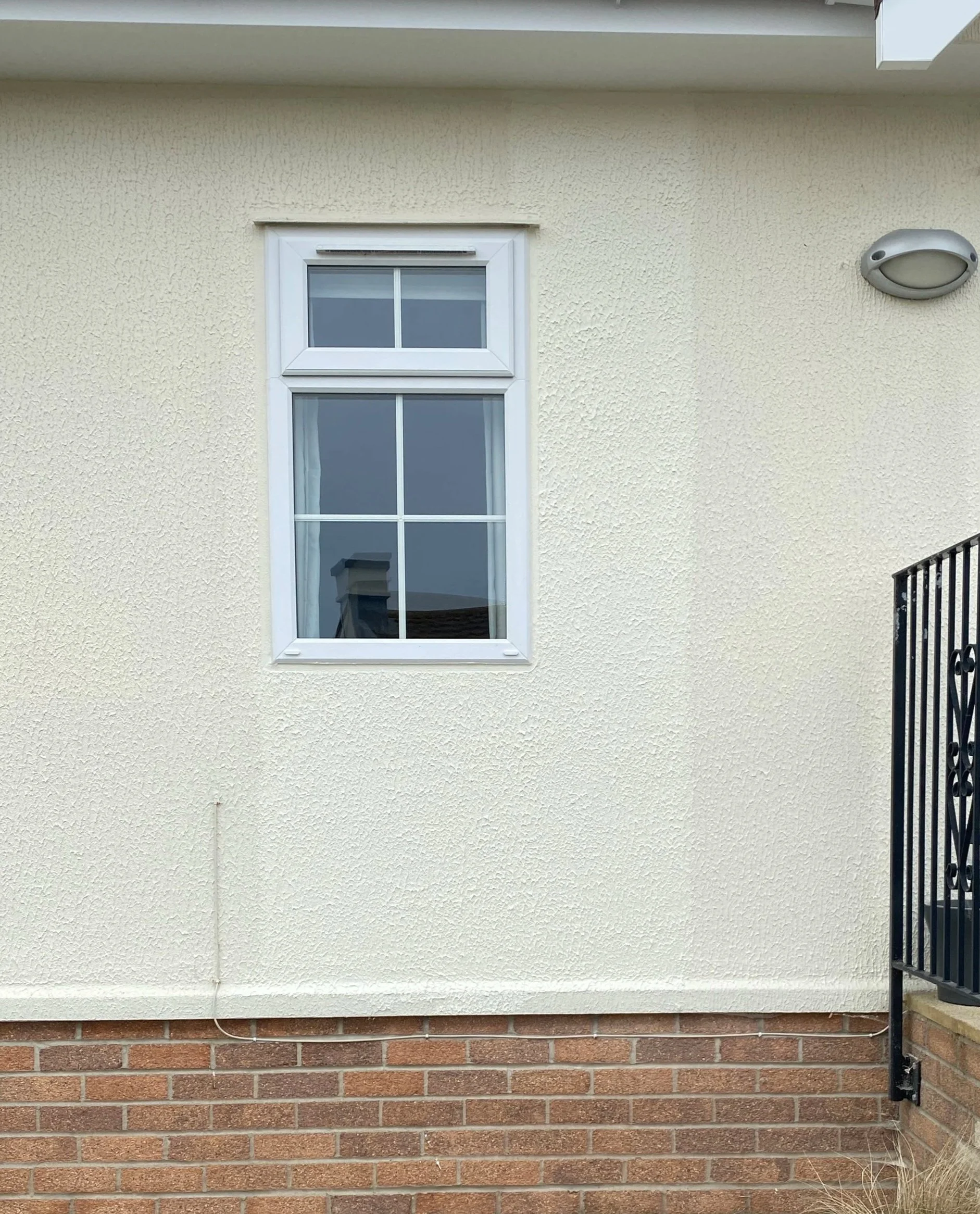 A close-up view of a white-framed double-pane window on a beige stucco wall, with a small portion of a black metal railing on the right side and a brick foundation at the bottom.