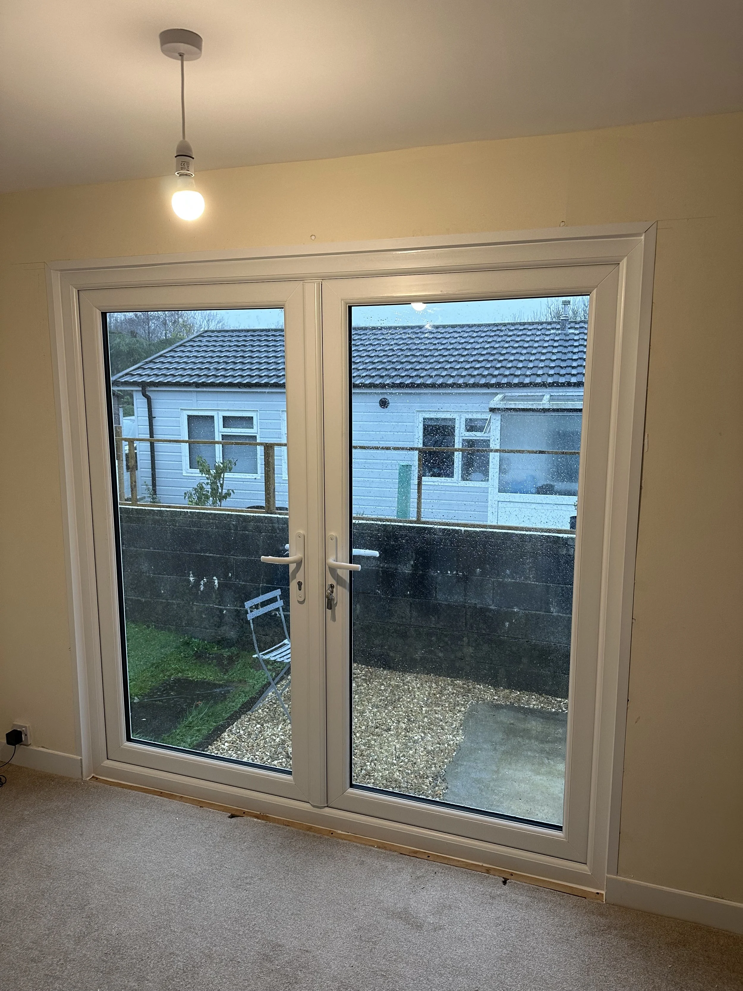 Interior view of a room with glass double doors leading outside, showing a small backyard with a chair, a gravel yard, and another house in the background.