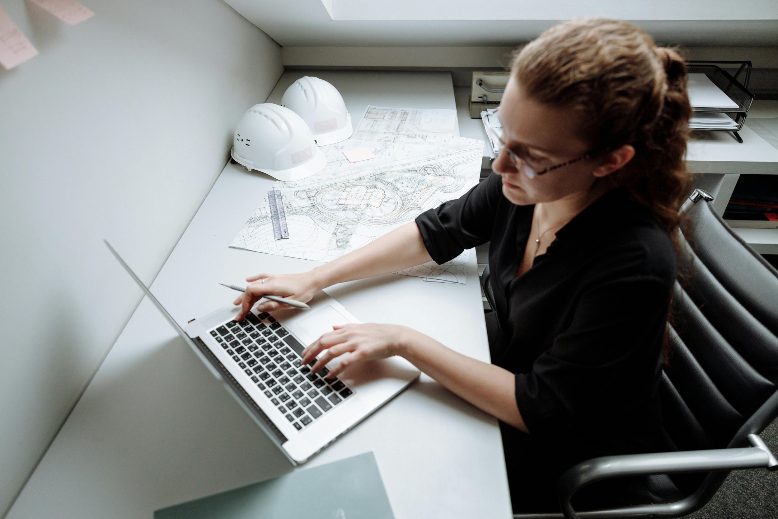 A woman works on a laptop at a desk with construction blueprints and two safety helmets beside her. Reflecting the perspective of Steven Jermoluk on shaping construction timelines.