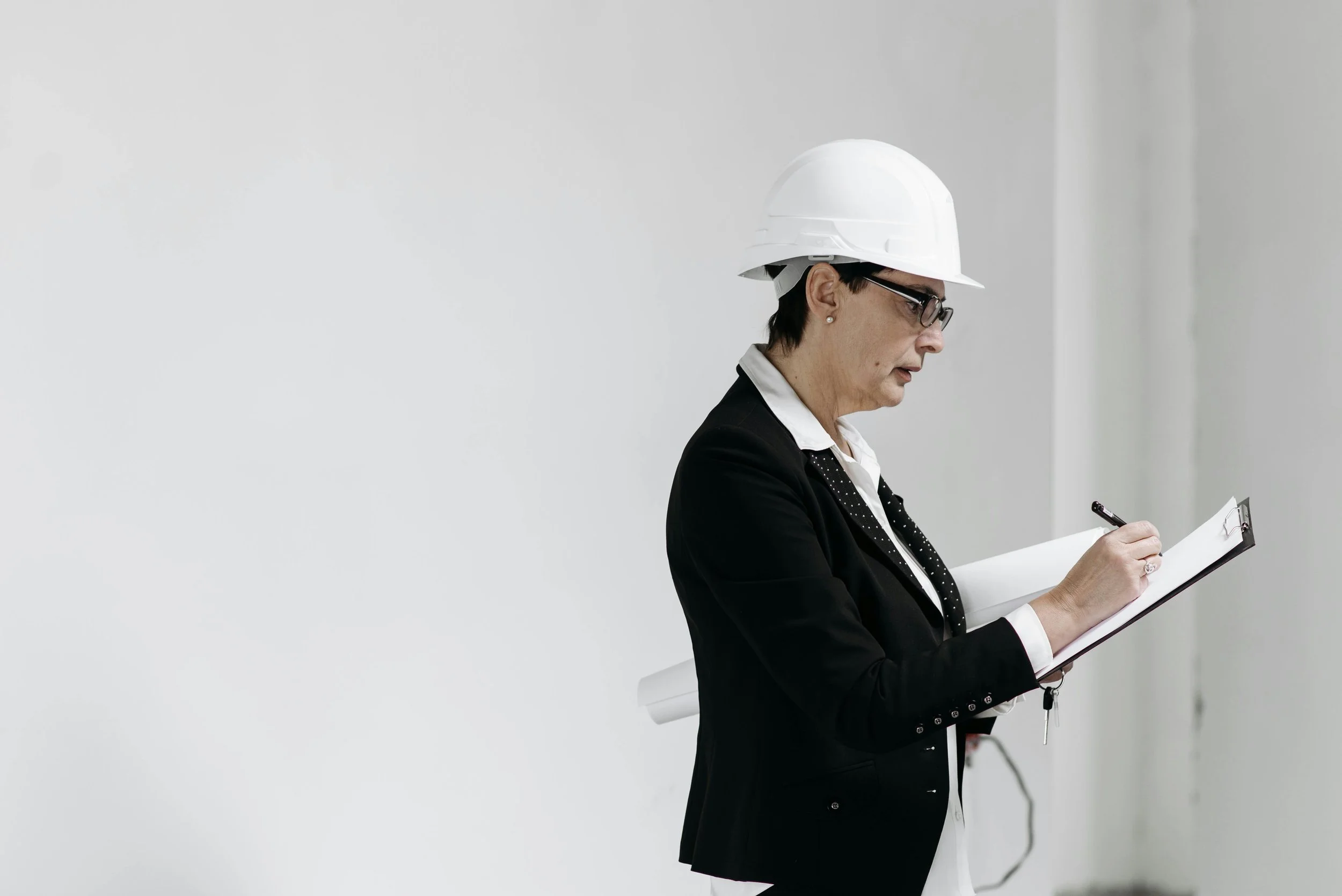 A woman in a black suit and white hard hat writes on a clipboard while inspecting a site. She appears focused, likely conducting a construction or safety evaluation. Reflecting the perspective of Steven Jermoluk on uncovering construction costs.