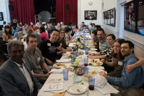 People gathered at a long banquet table with food and drinks, participating in a group meal in a large room with white walls and framed pictures.