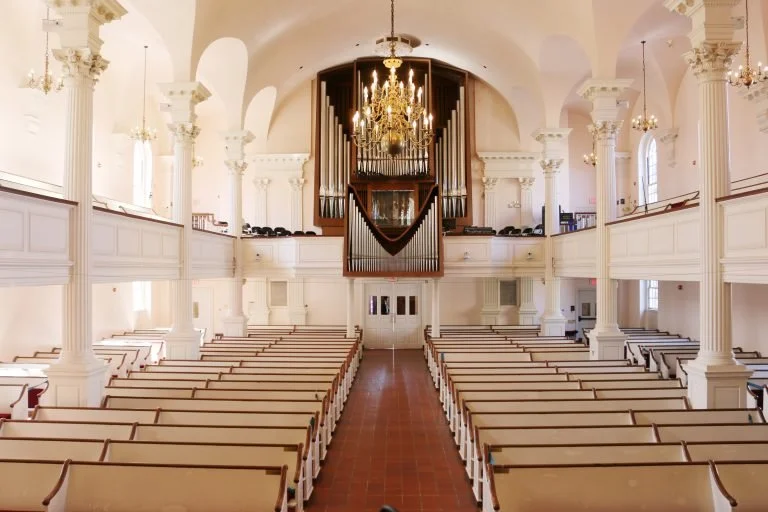 Interior of a church with white walls, wooden pews, large arched windows, and a pipe organ with a chandelier above.