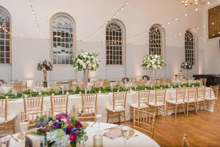 Wedding reception setup with long table, gold chairs, floral centerpieces, string lights, and a white wall with arched windows.