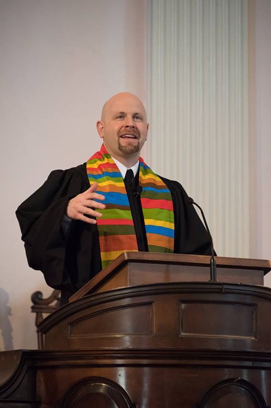A man in a black robe giving a speech at a wooden podium, wearing a colorful rainbow stole.