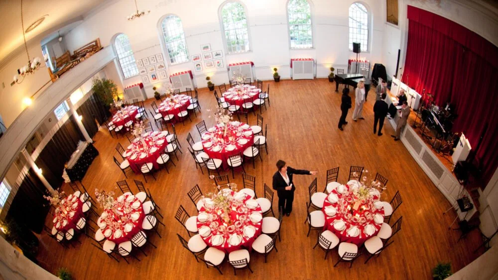Banquet hall with round tables covered with red tablecloths and white chairs, set for a formal event with floral centerpieces, a piano, and a small stage area with a red curtain.