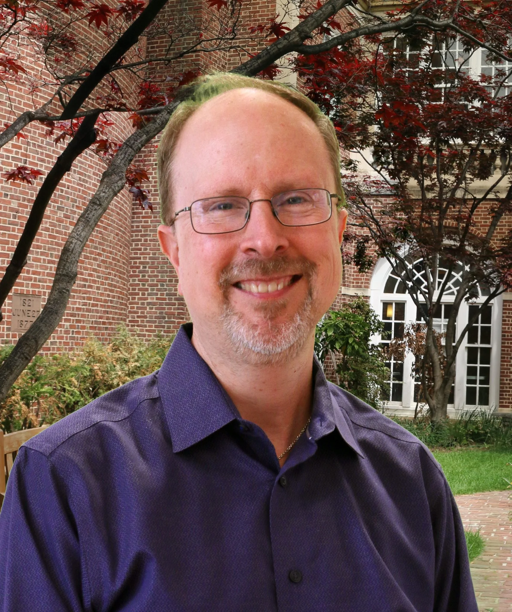 A smiling man with glasses and a goatee, wearing a purple shirt, is outdoors in front of a brick building with a garden and trees.