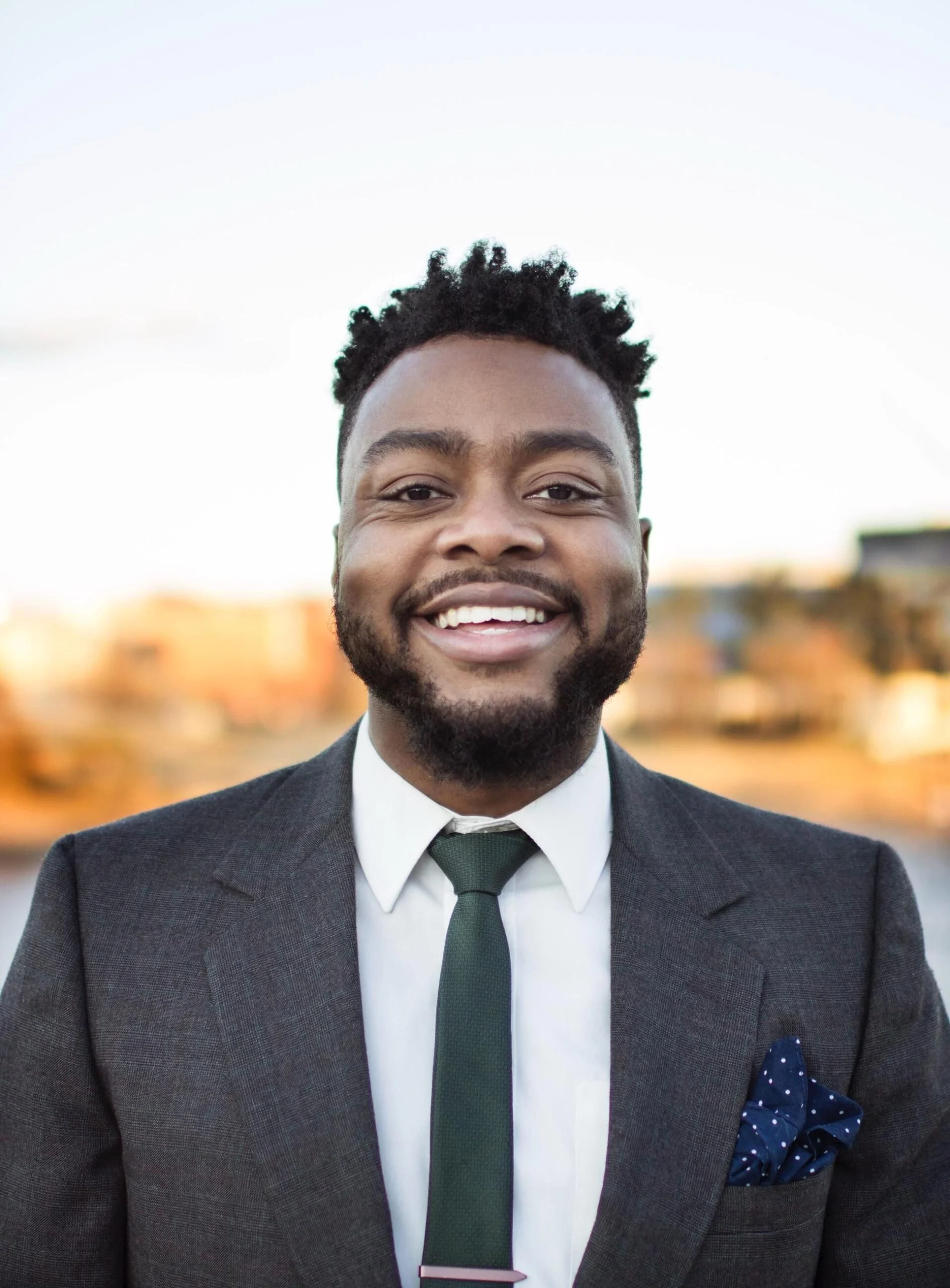 Portrait of a smiling Black man wearing a dark suit, white shirt, green tie, and pocket square, outdoors in front of a blurred cityscape.