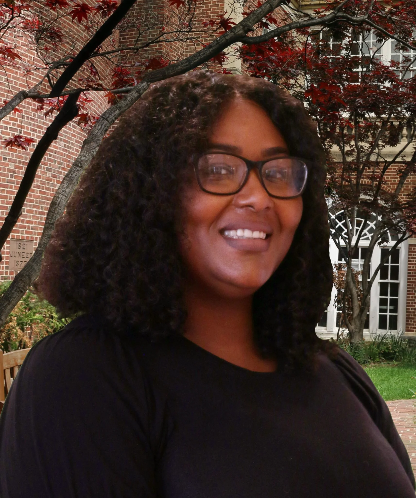 A woman smiling outdoors in front of a brick building with trees and greenery.