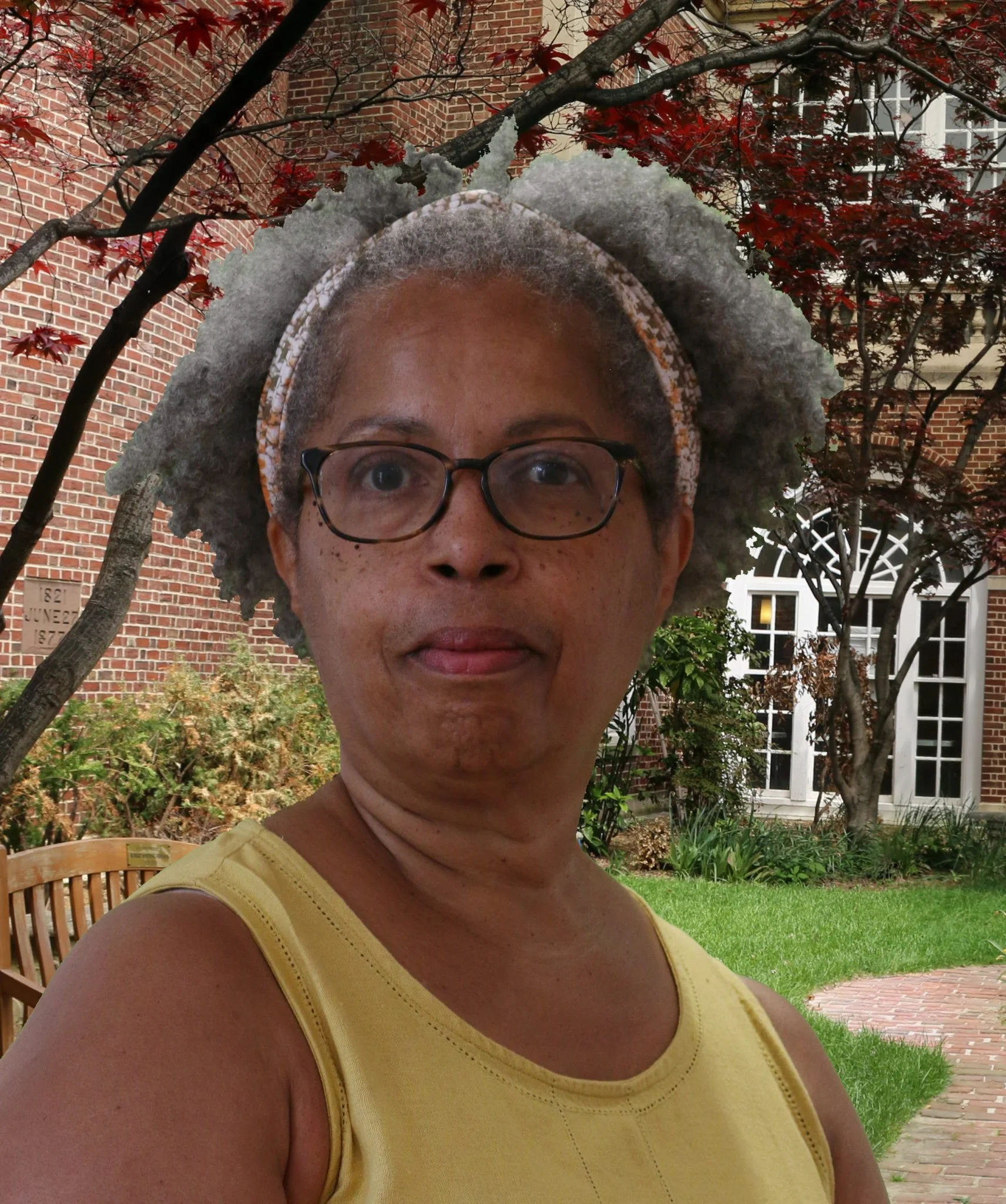A woman with gray, curly hair, glasses, wearing a yellow sleeveless top, standing outdoors in front of a brick building with trees and green grass.