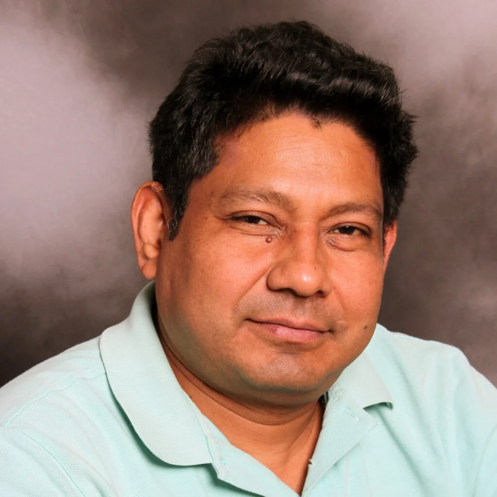 Close-up portrait of a man with dark hair, wearing a light green polo shirt, against a dark blurred background.