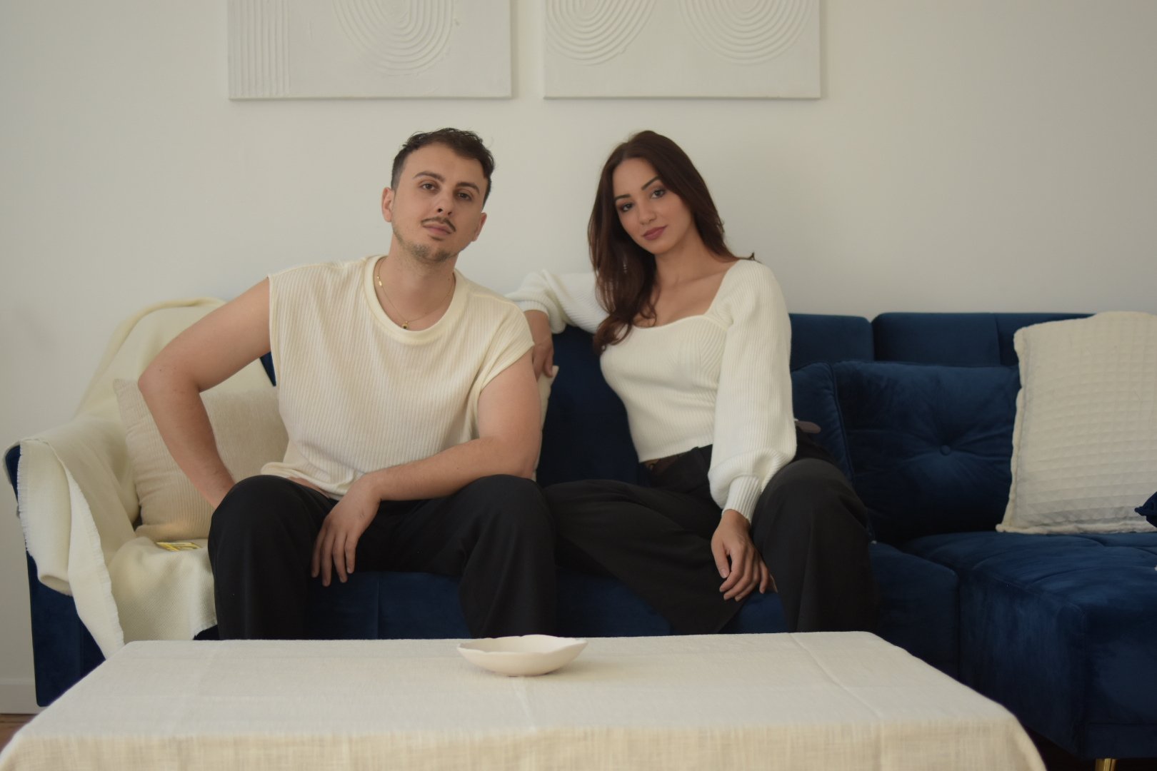 Deux jeunes adultes, un homme et une femme, assis sur un canapé bleu dans un salon, regardent l'objectif.