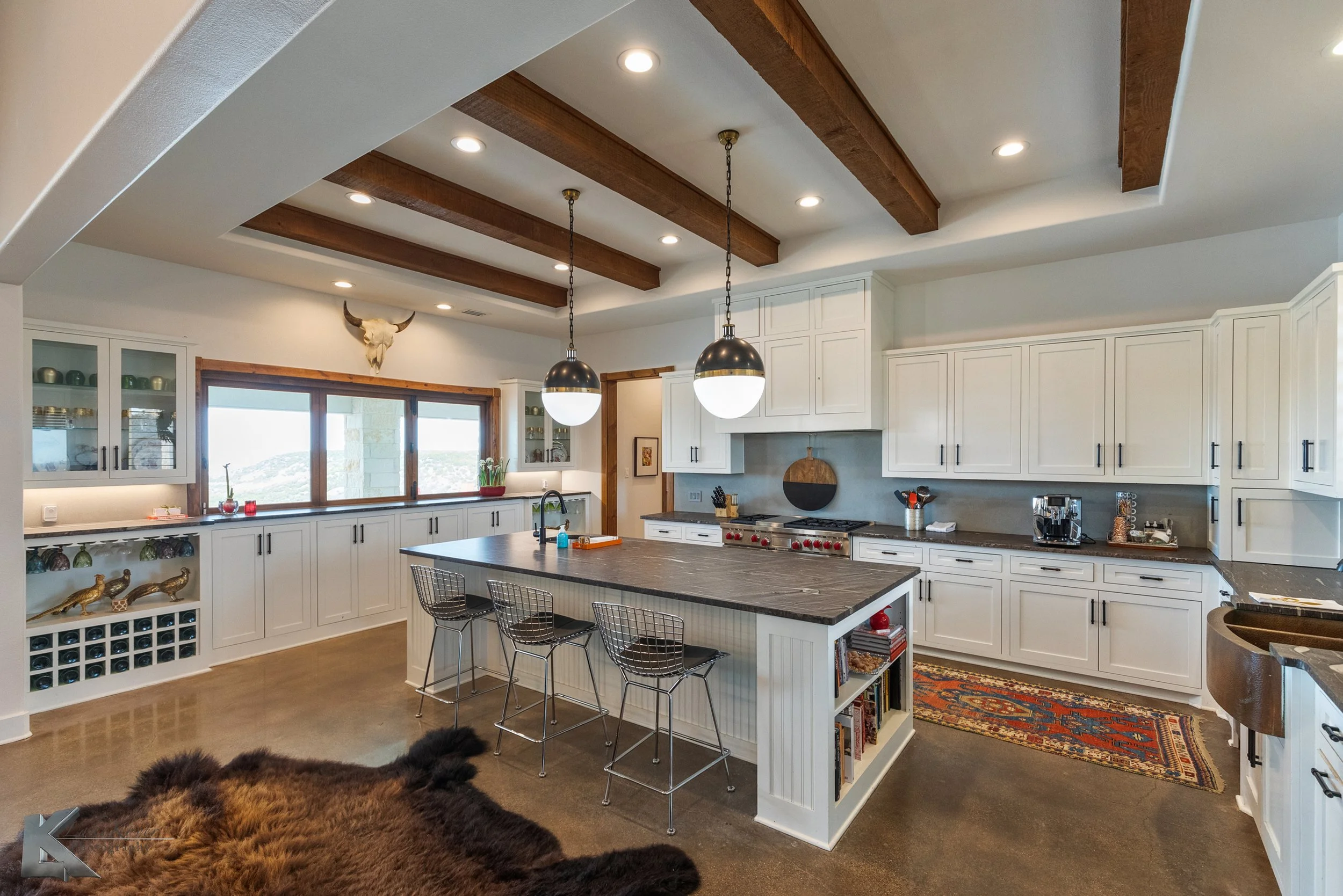 The kitchen has wood beam accents, a massive island with built-in shelving, dual sinks, & stone countertops.